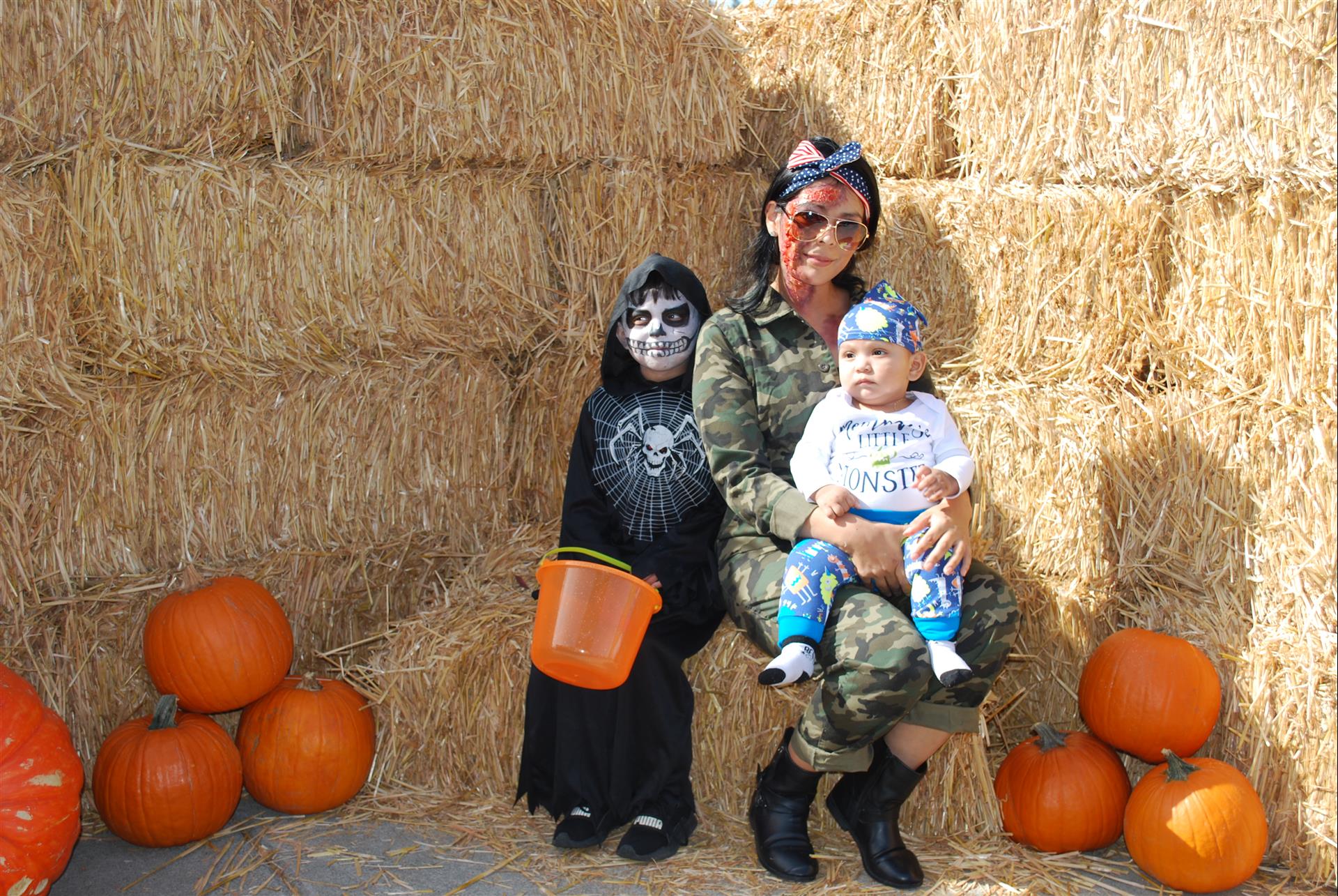 A woman and two children are sitting on a bale of hay surrounded by pumpkins.