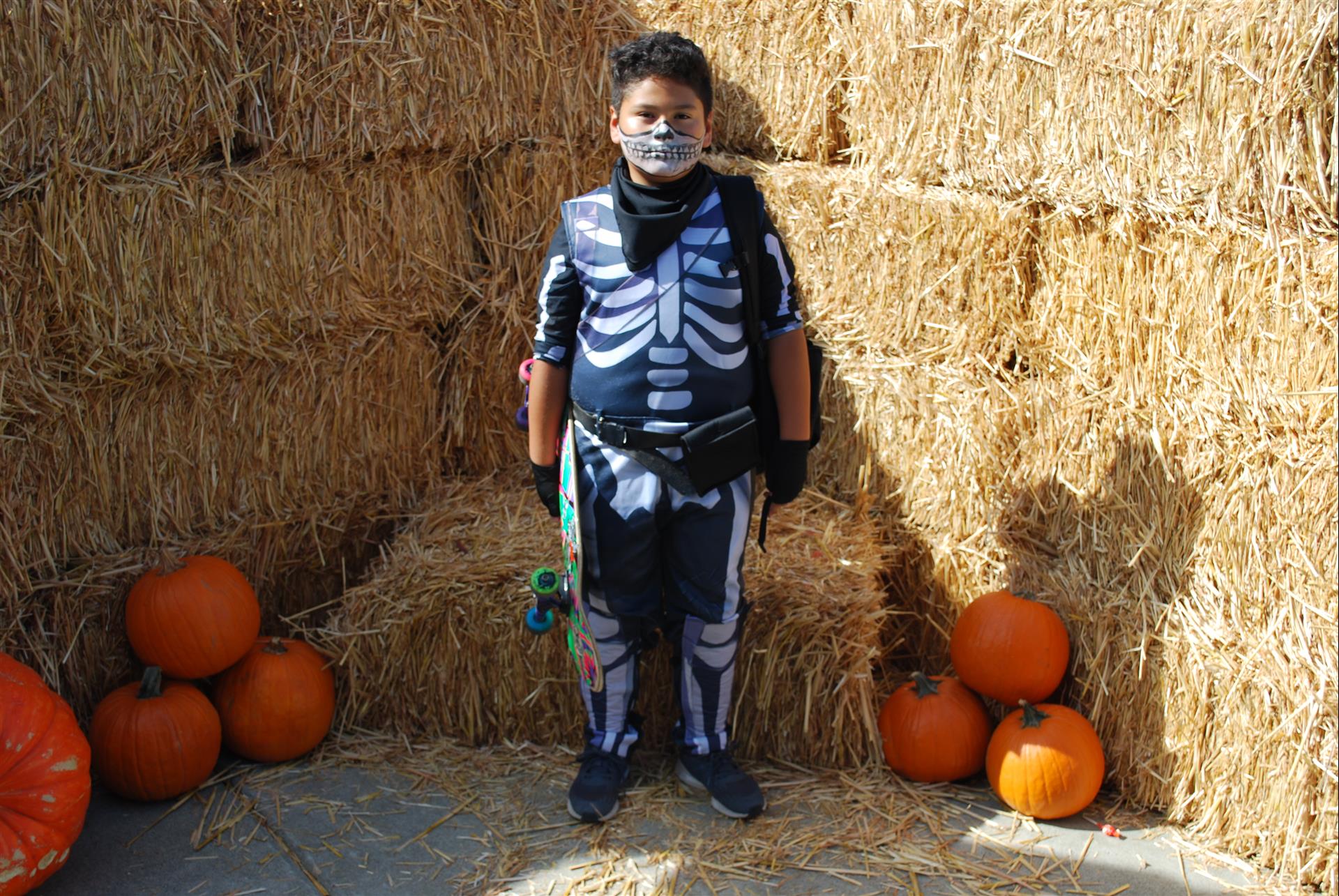 A boy in a skeleton costume is standing in front of hay bales and pumpkins.