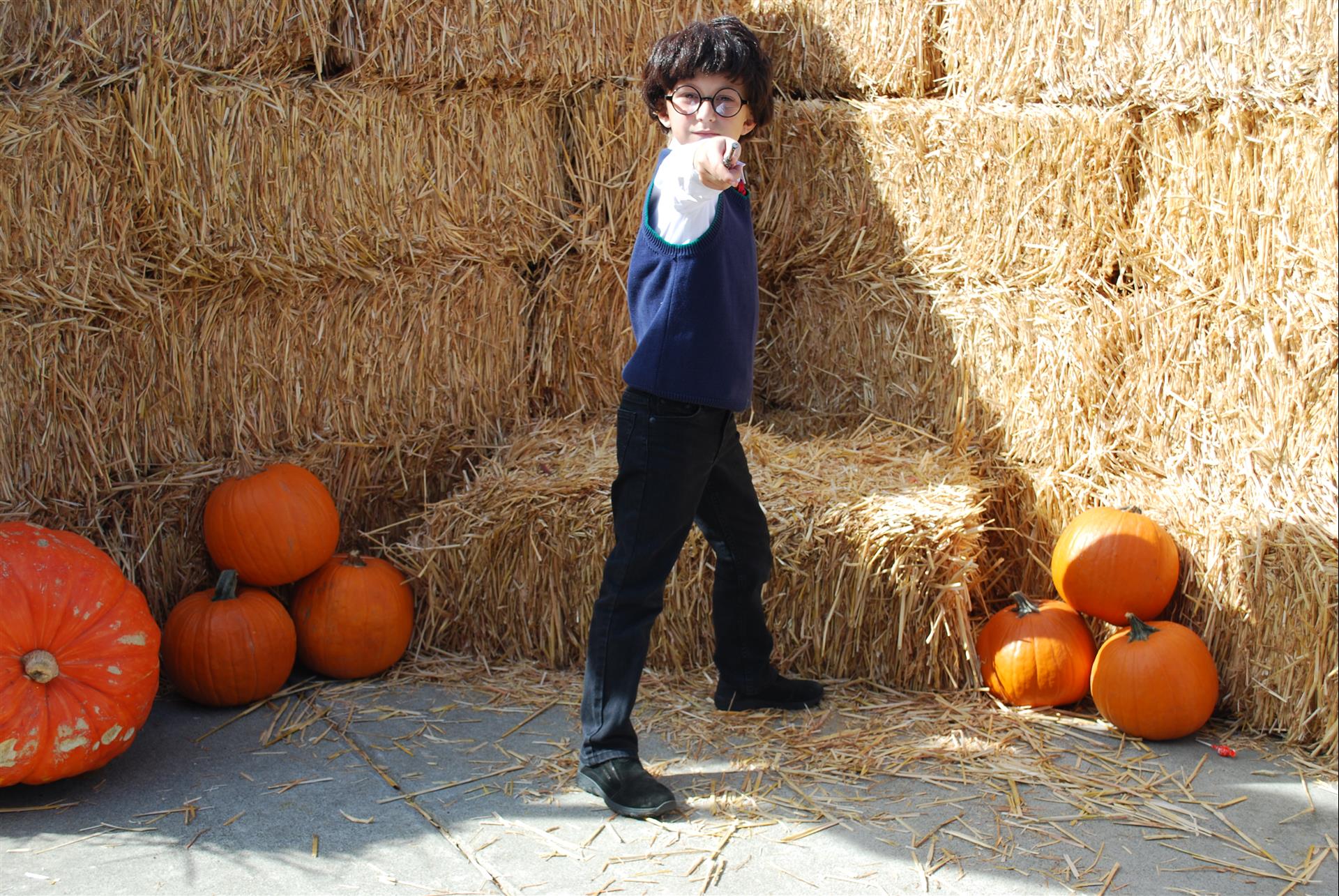 A young boy is pointing at the camera in front of a pile of hay bales and pumpkins.