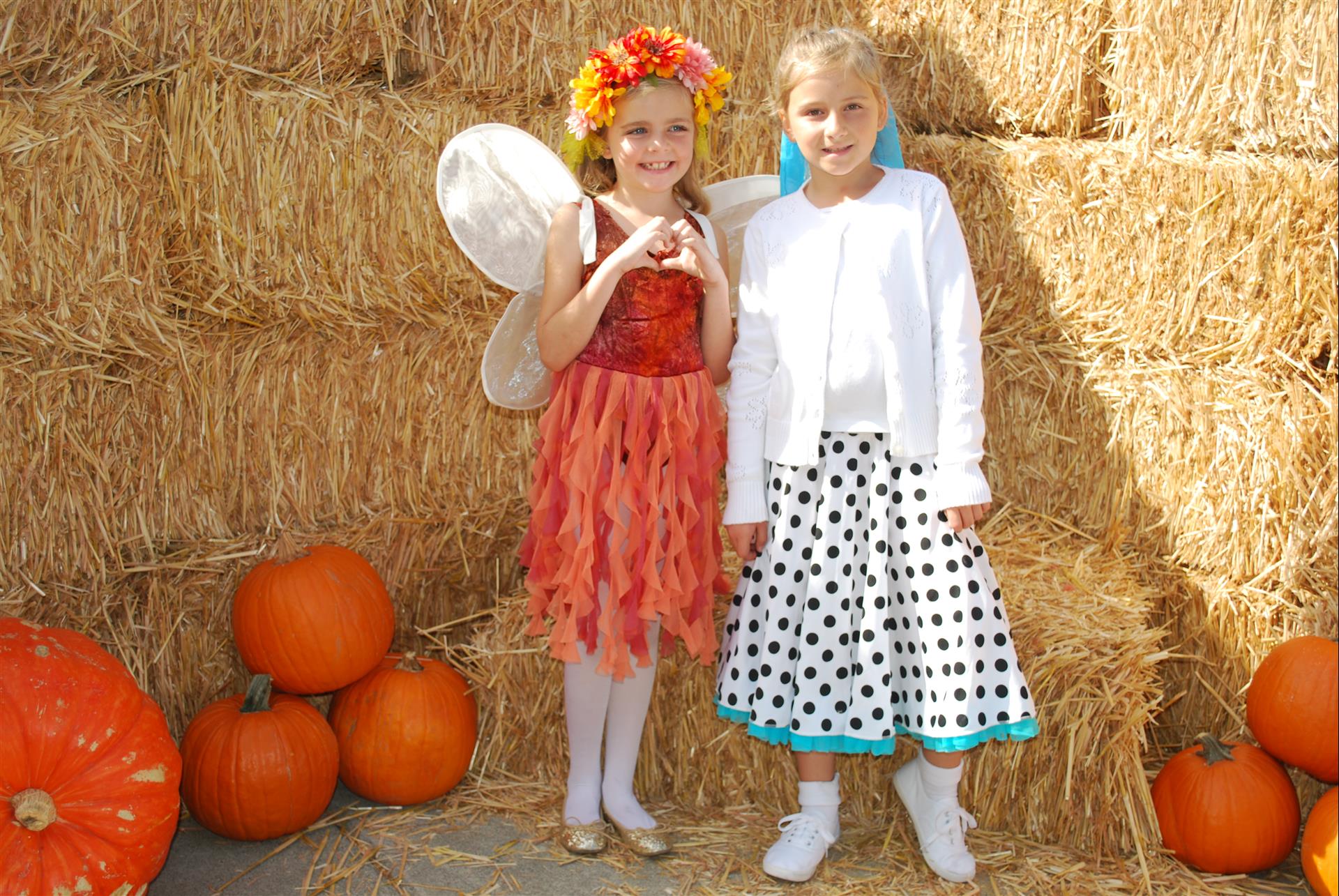 Two little girls in costumes are standing in front of hay bales and pumpkins.