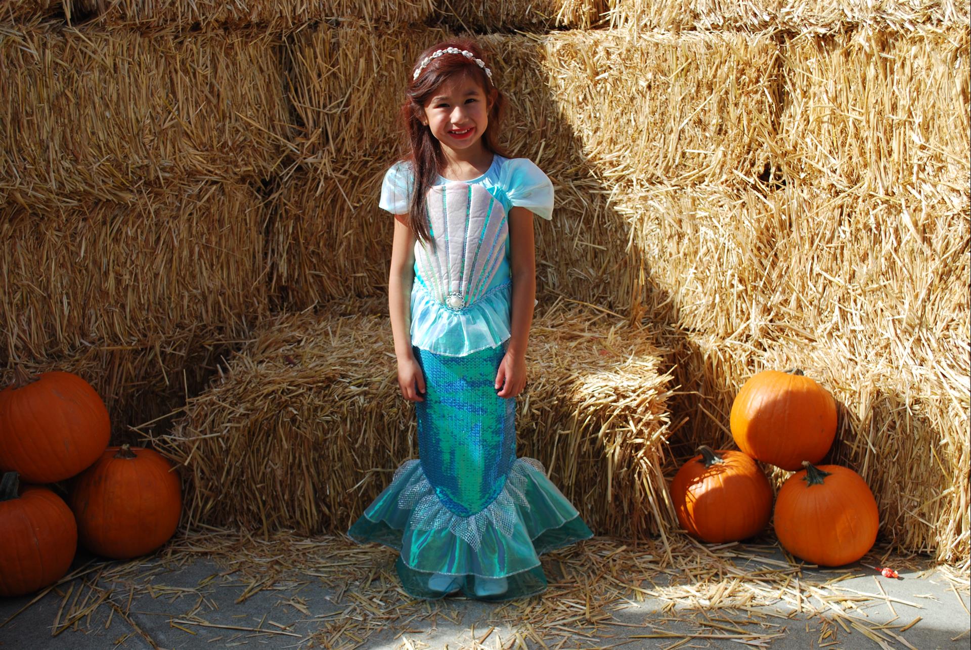 A little girl in a mermaid costume is standing in front of hay bales and pumpkins.