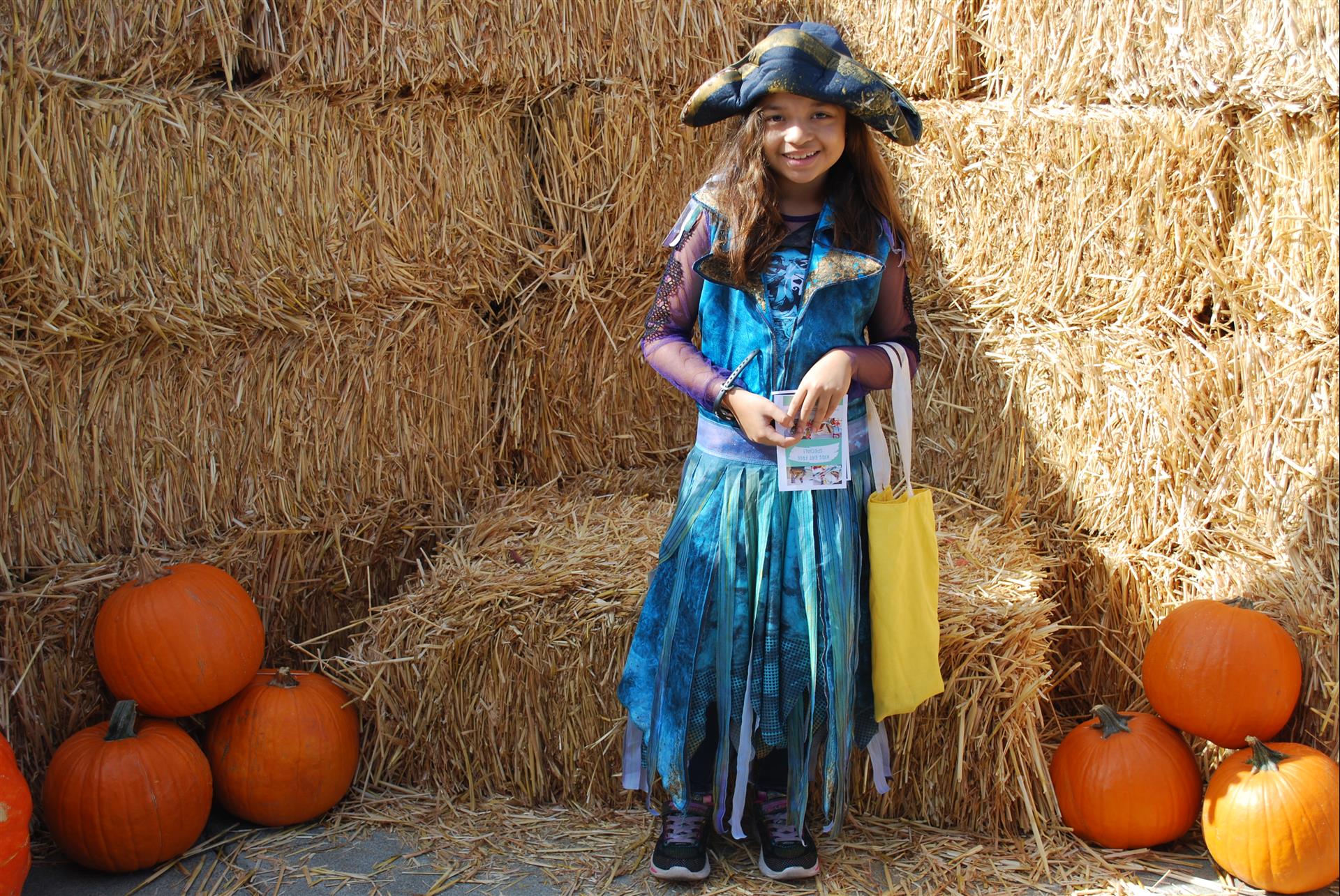 A little girl in a pirate costume is standing in front of hay bales and pumpkins.