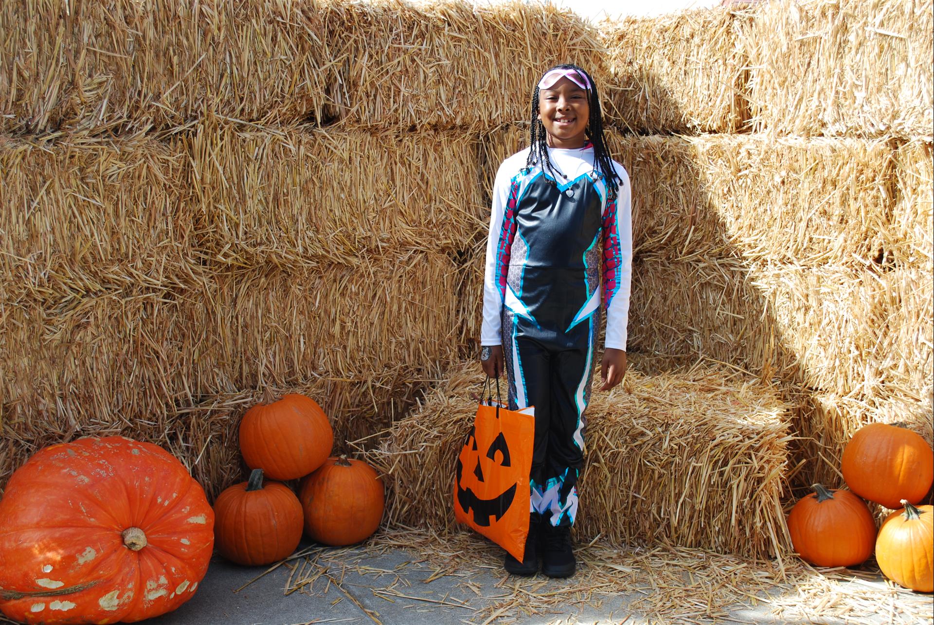 A girl in a halloween costume is standing in front of hay bales and pumpkins