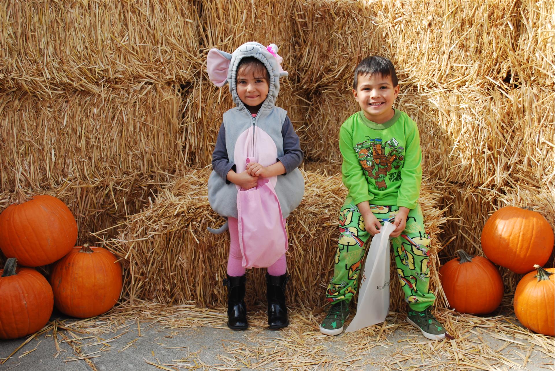 A boy and a girl in costumes are sitting on hay bales surrounded by pumpkins.