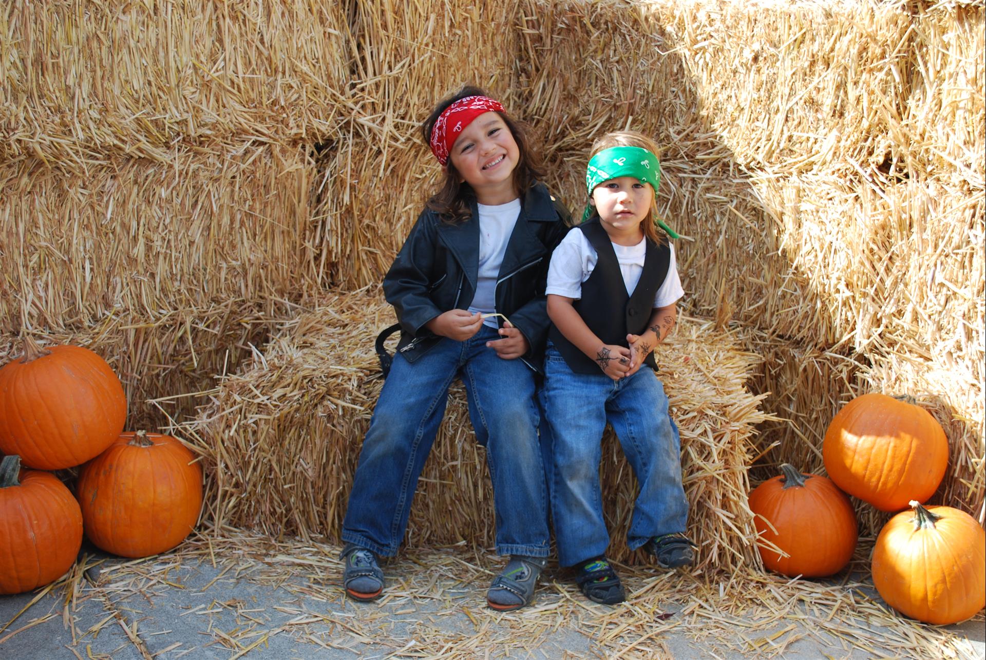 Two children are sitting on a bale of hay surrounded by pumpkins.