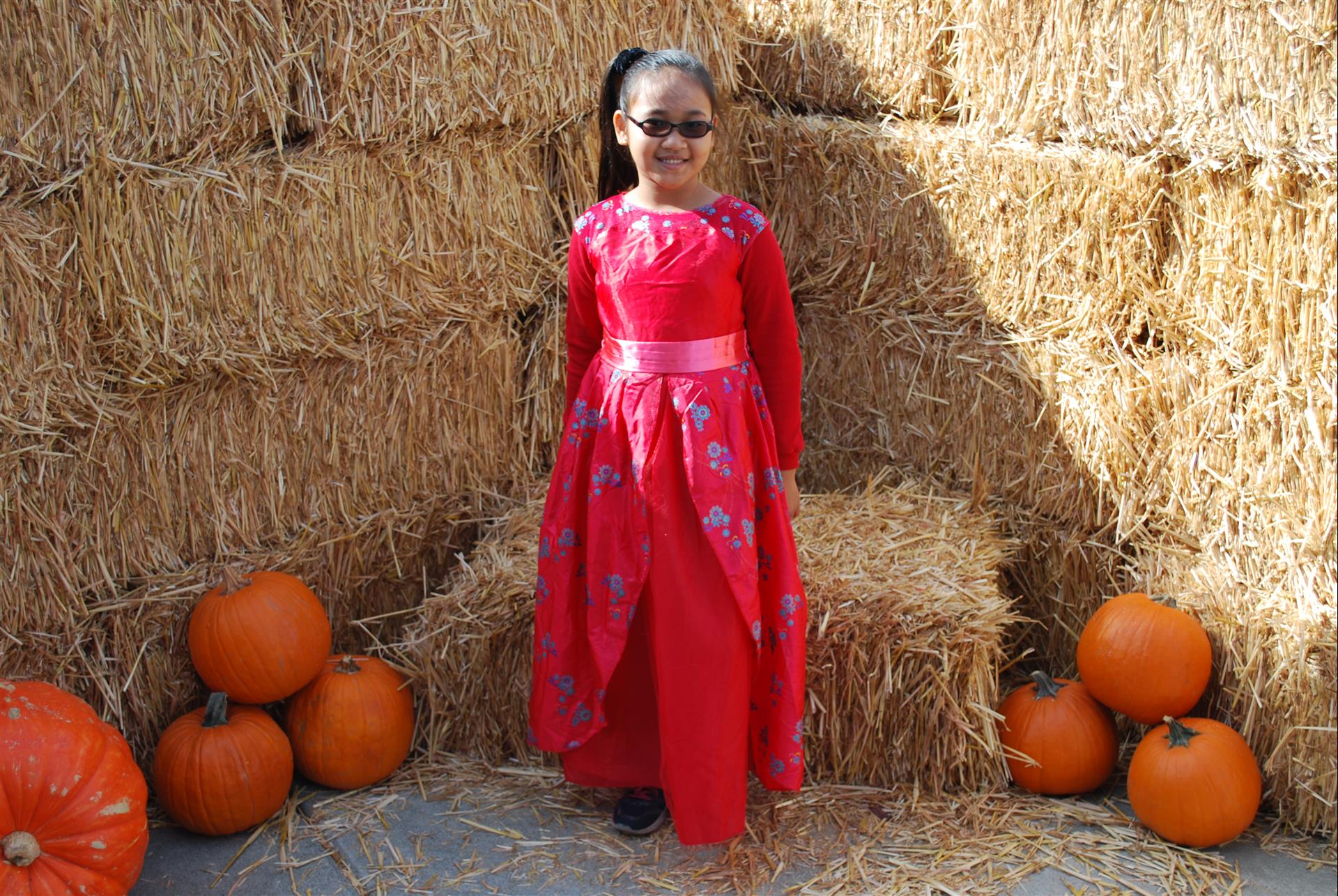 A little girl in a red dress is standing in front of hay bales and pumpkins.