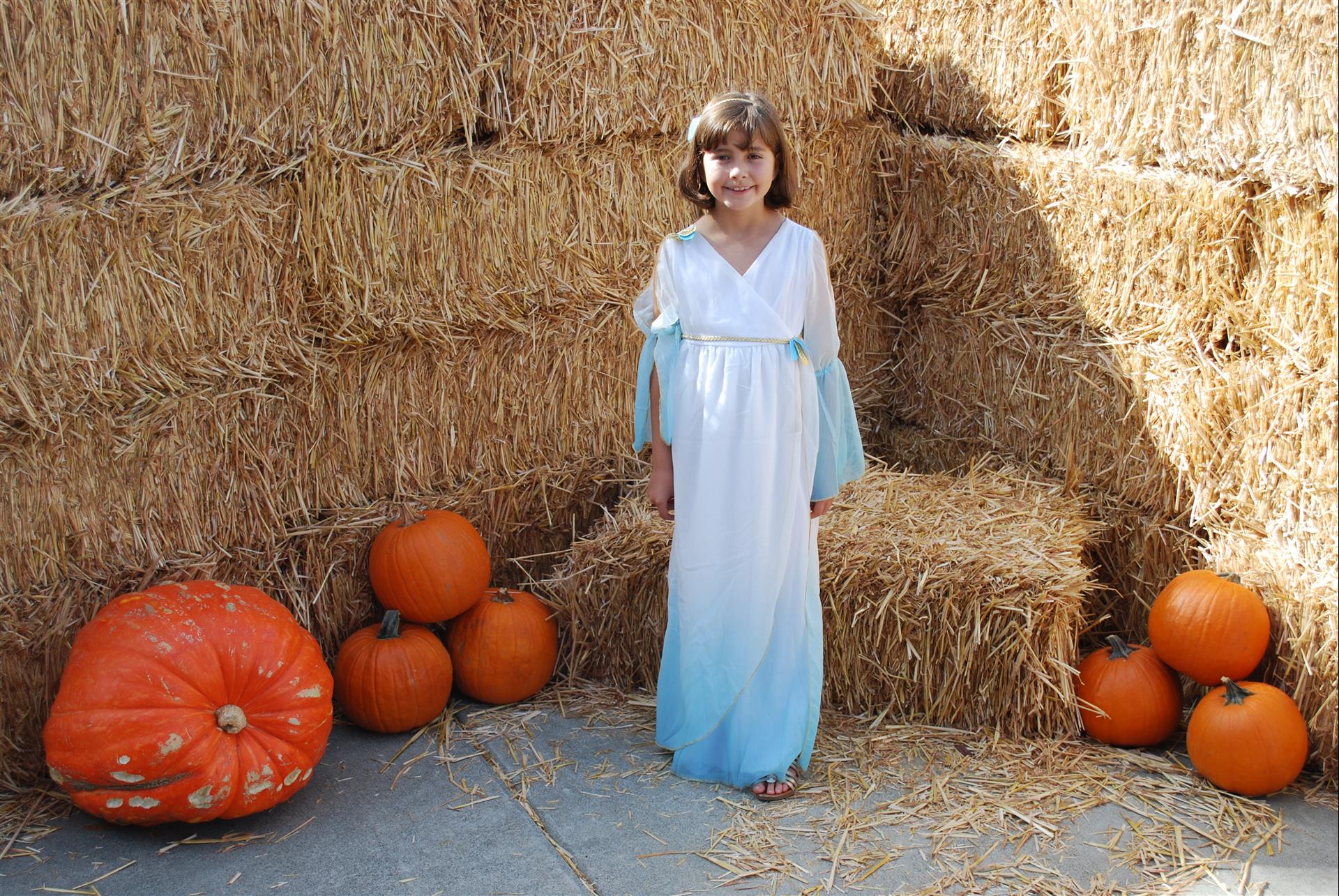A little girl in a white dress is standing in front of hay bales and pumpkins.