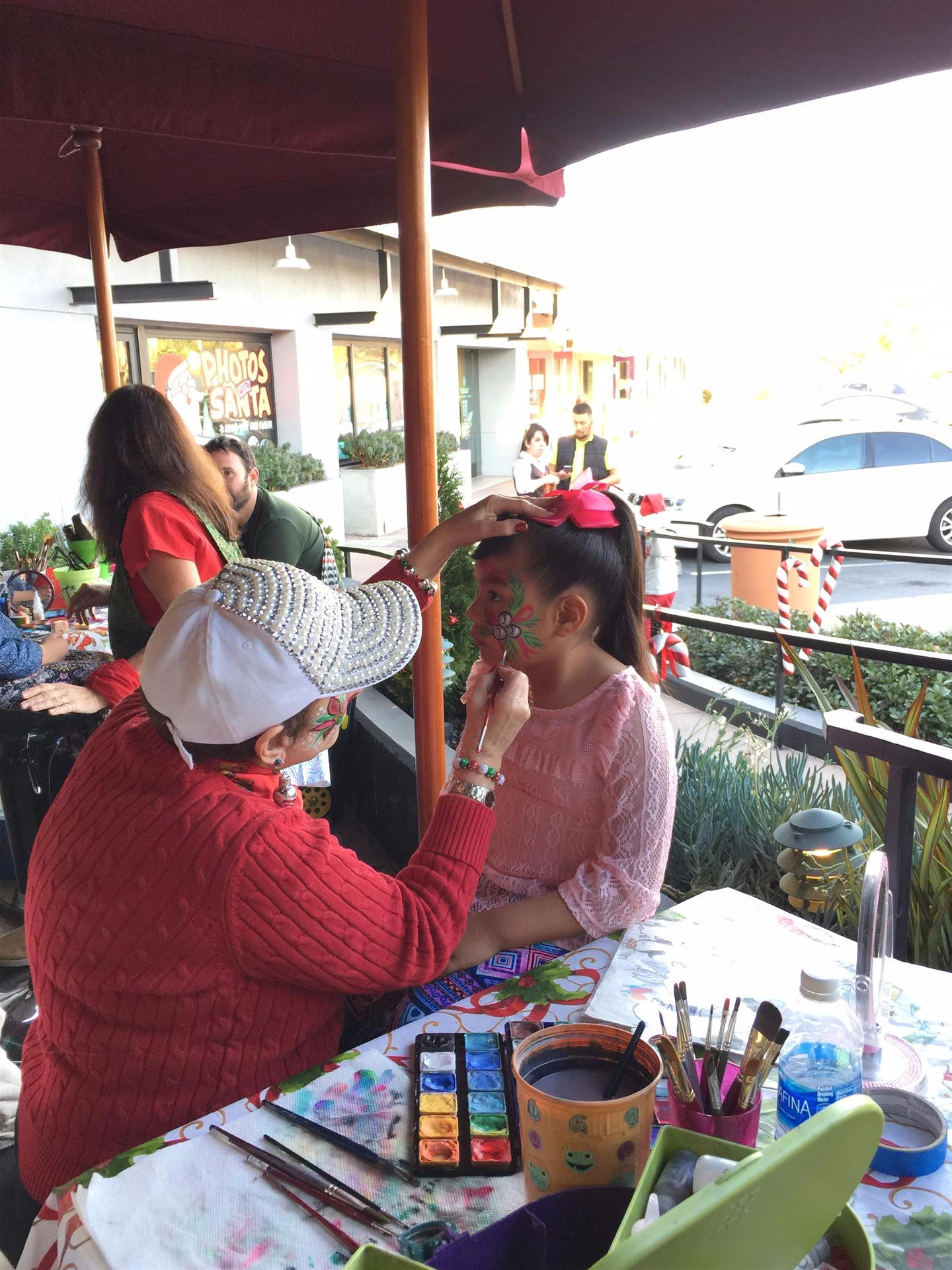 A woman is painting a little girl 's face under an umbrella.