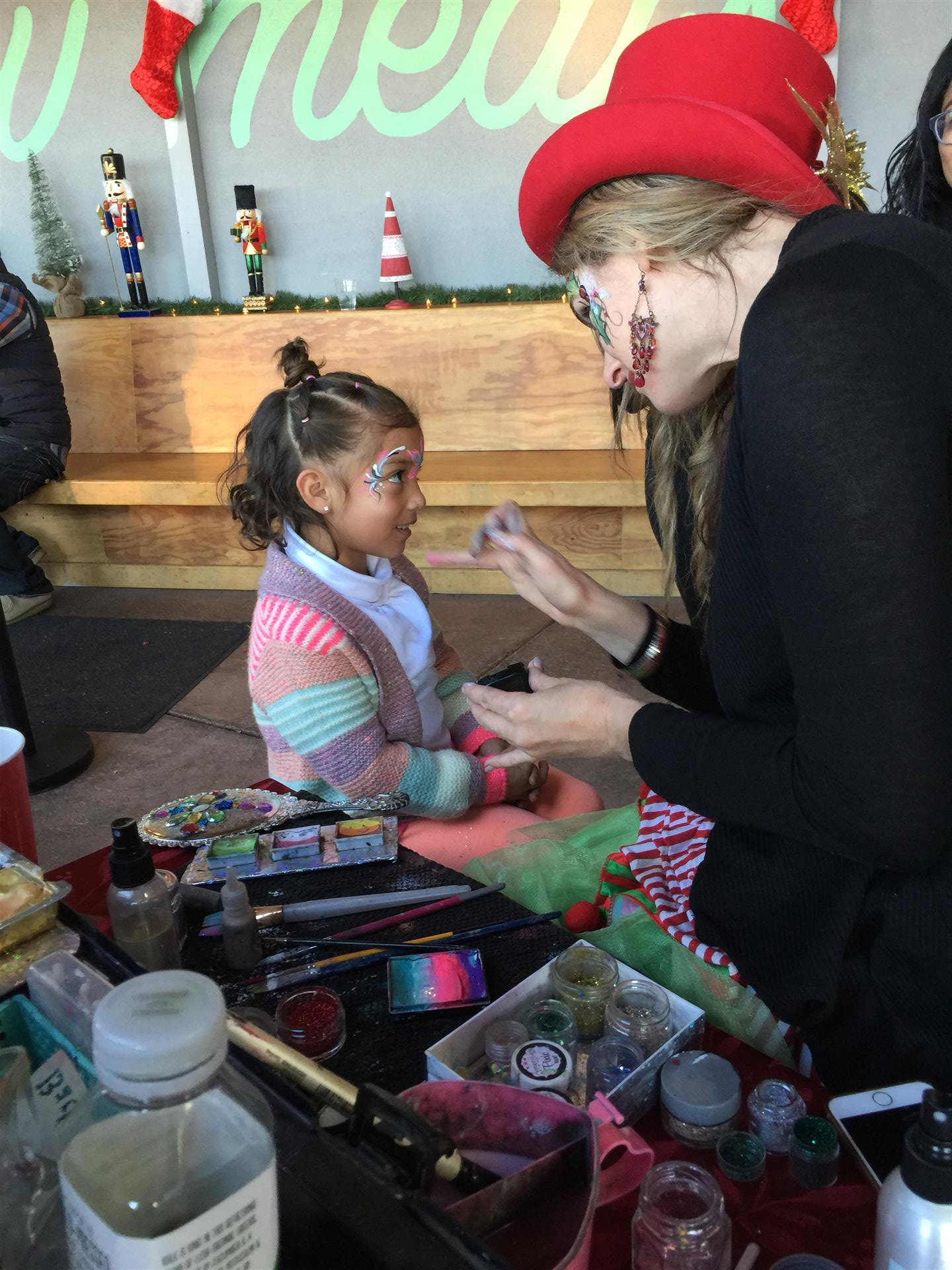 A little girl is getting her face painted by a woman in a red hat.