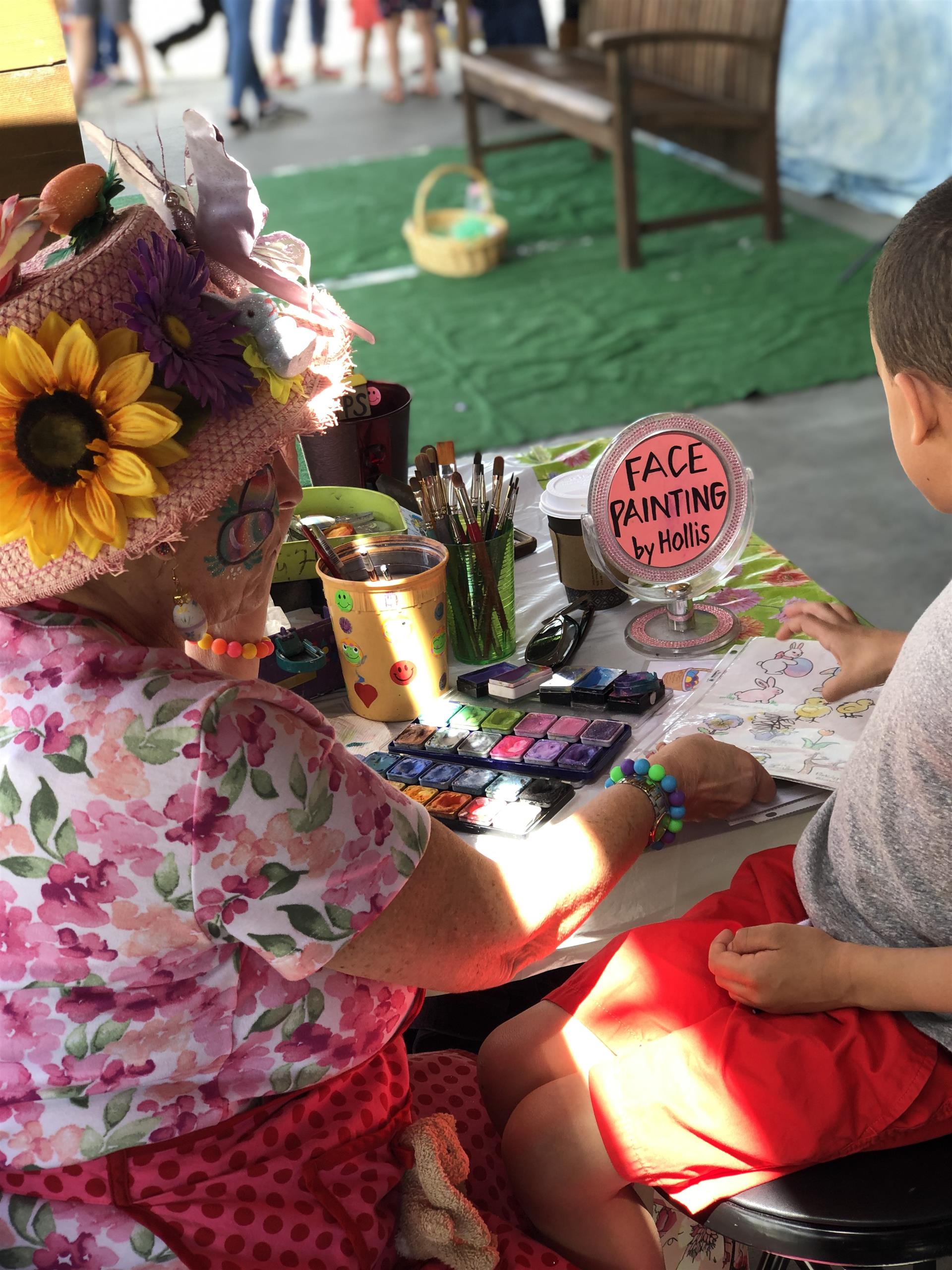 A woman is sitting at a table painting a child 's face.
