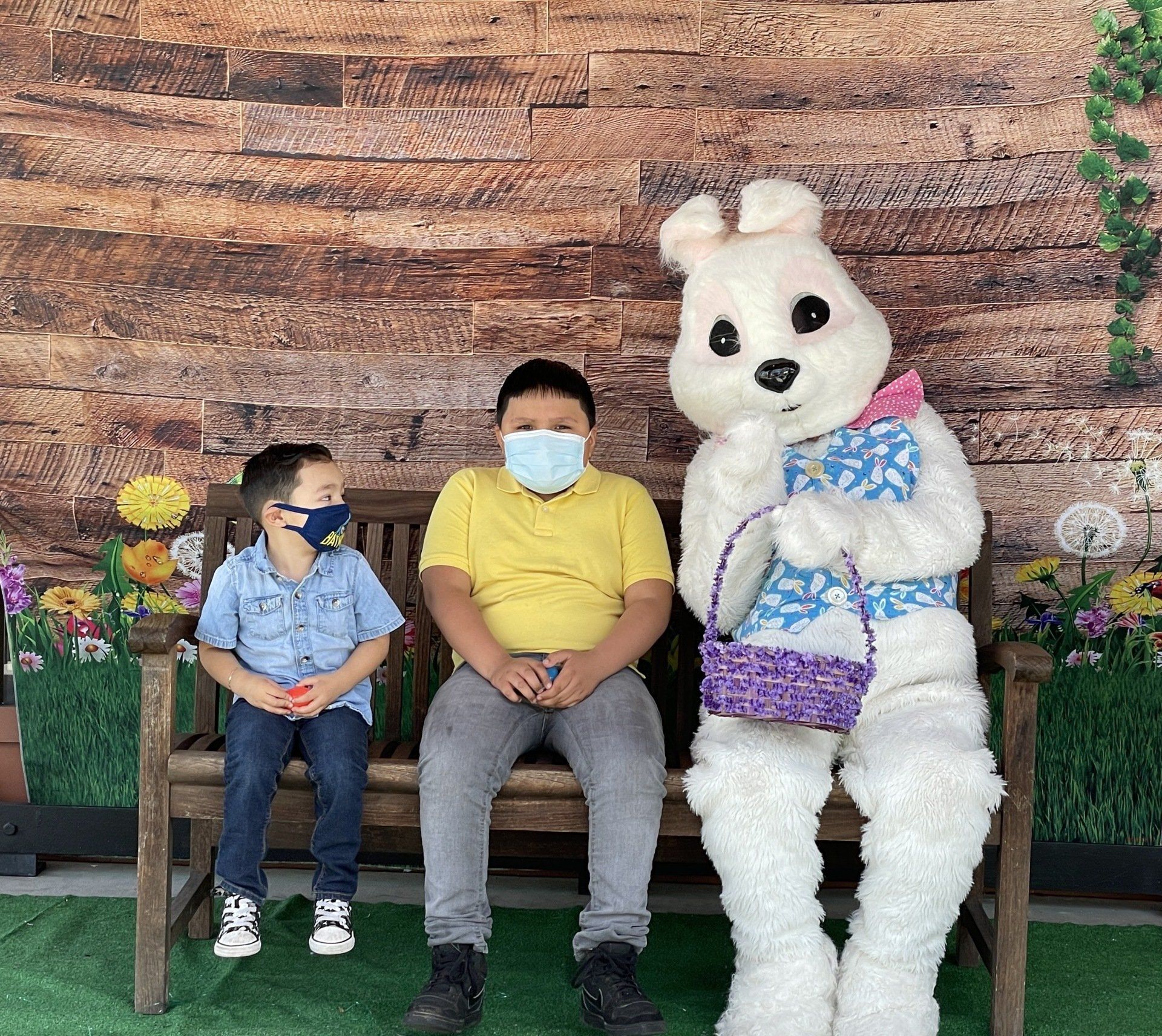 Two children wearing face masks are sitting on a bench next to an easter bunny.