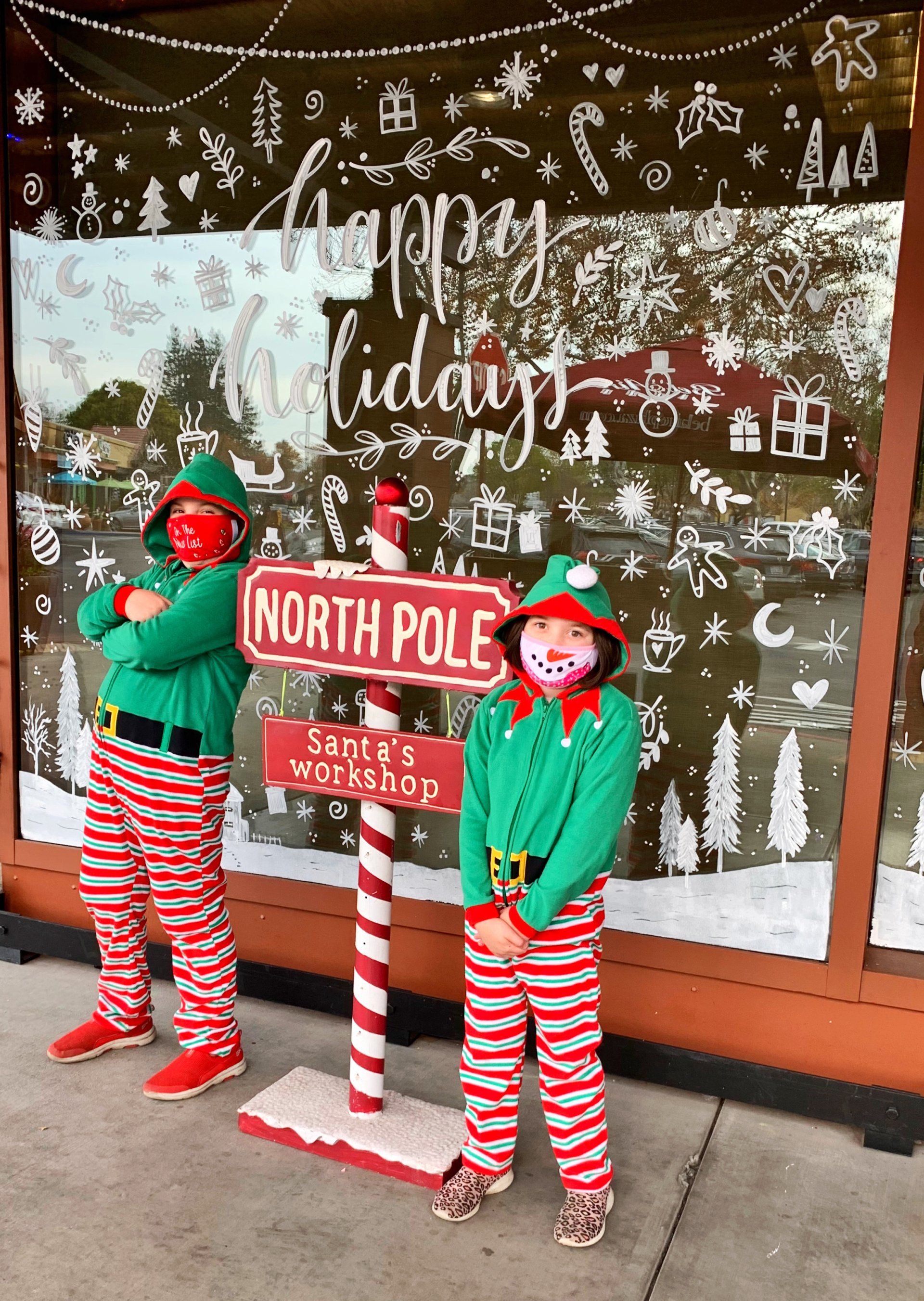 Two children dressed as elves are standing in front of a north pole sign.