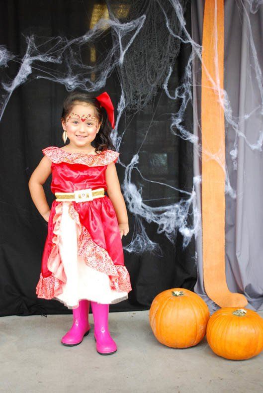 A little girl in a red dress and pink boots is standing next to pumpkins.