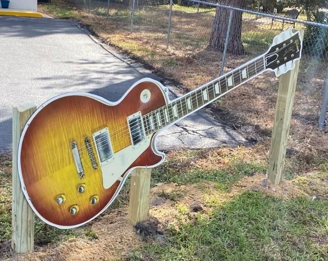 A large guitar is sitting on a wooden post in the grass