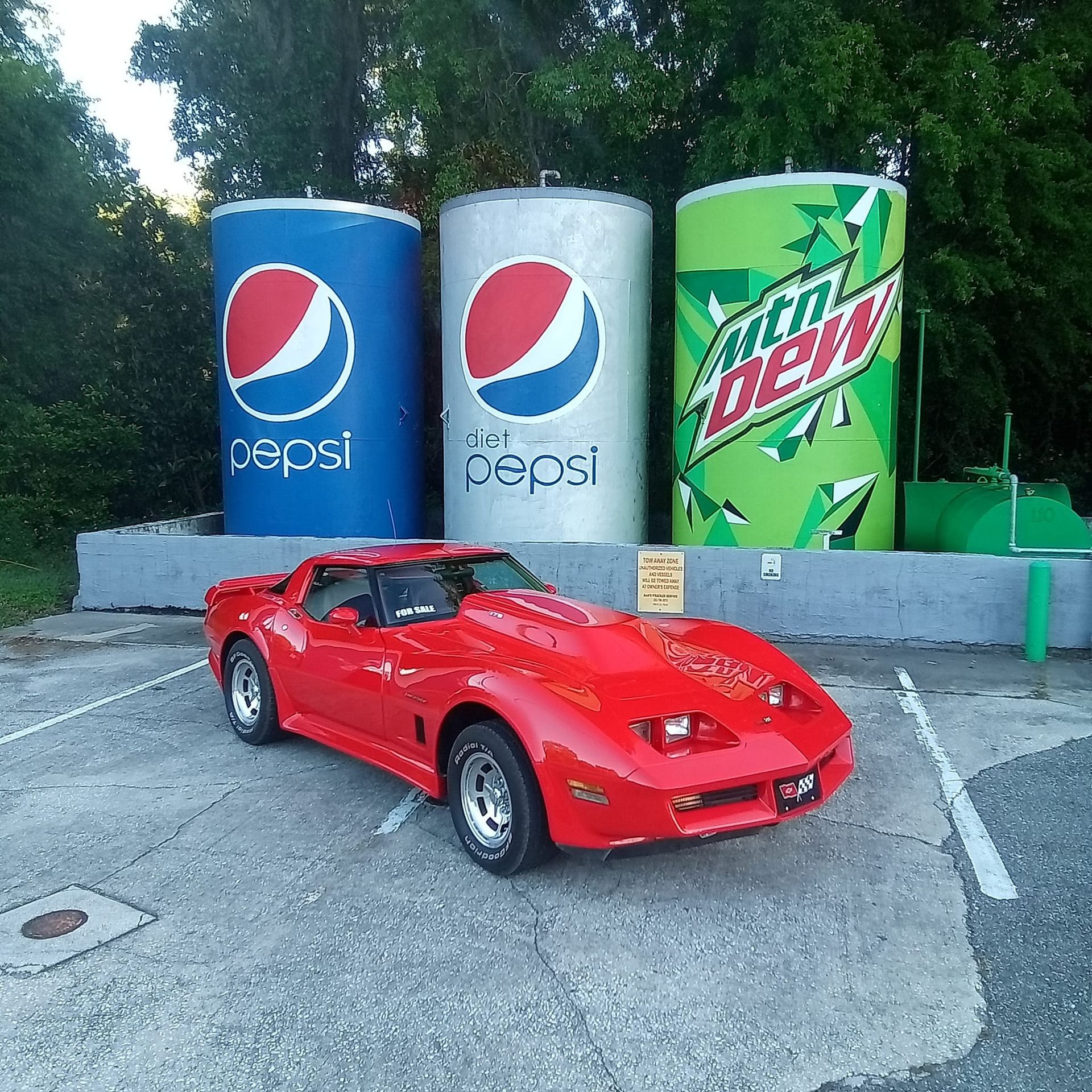 A red car is parked in front of giant pepsi and mountain dew cans
