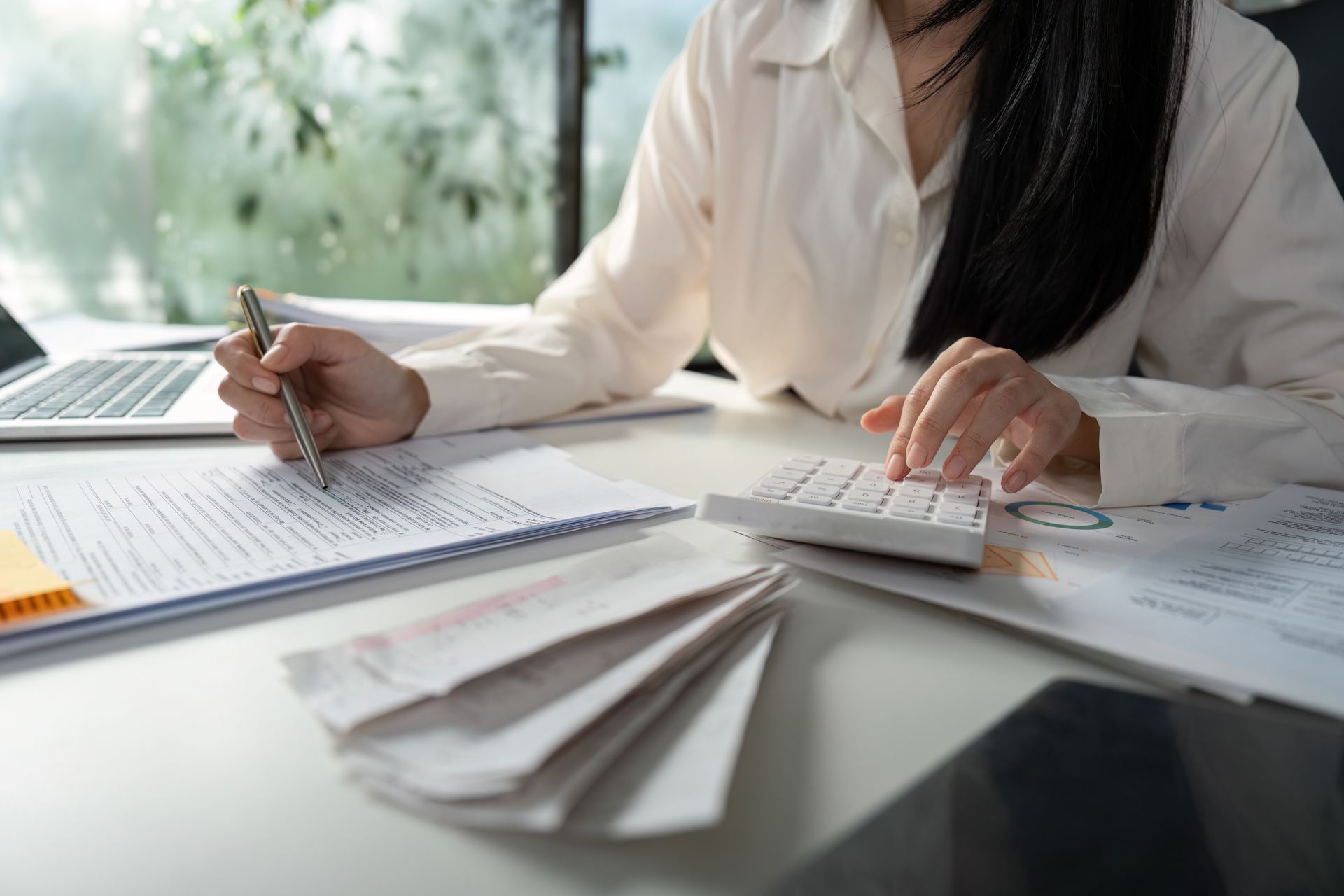 Woman in white shirt using a calculator, looking at documents with receipts and a laptop.