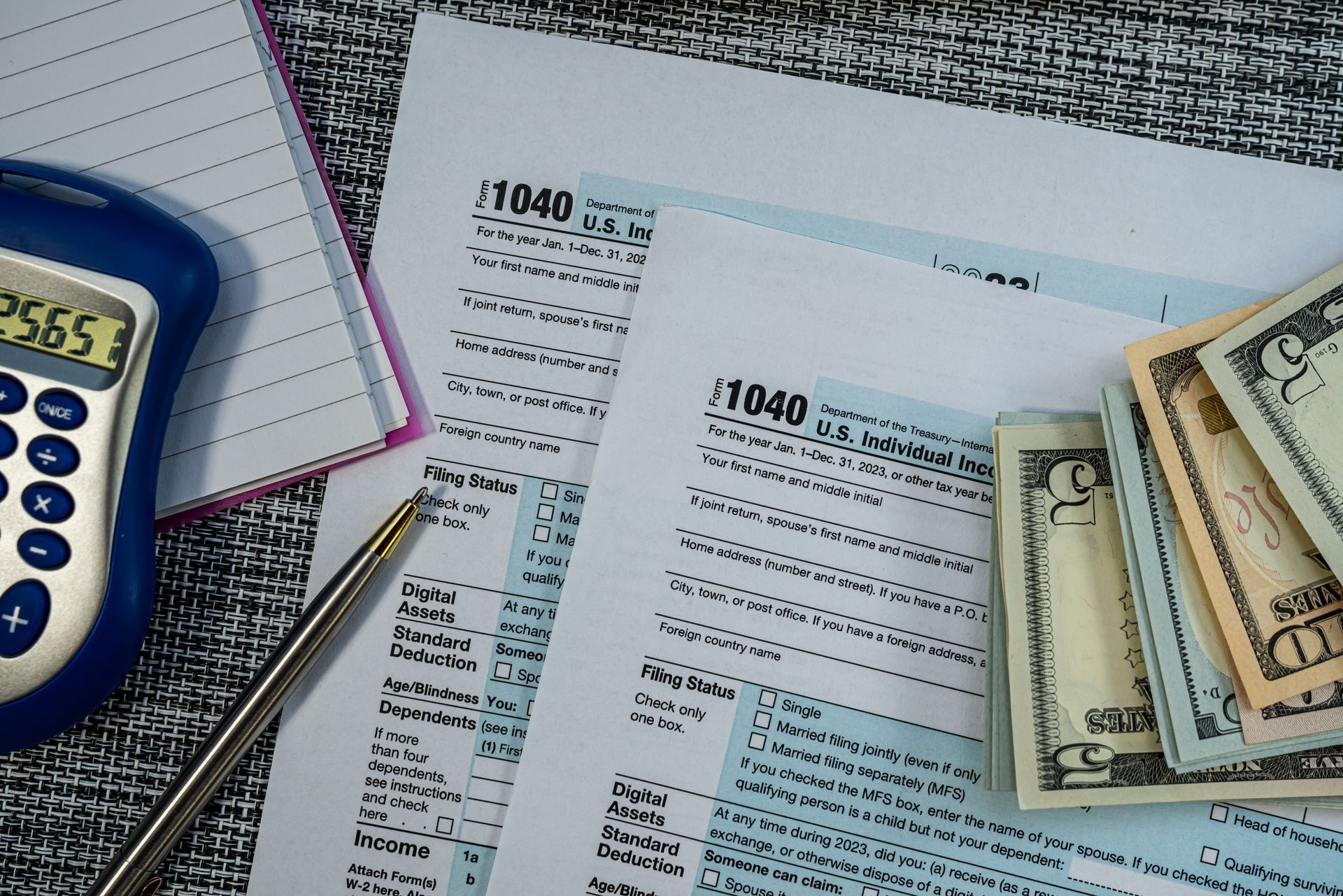 Tax forms, calculator, pen, notepad, and cash on a table.