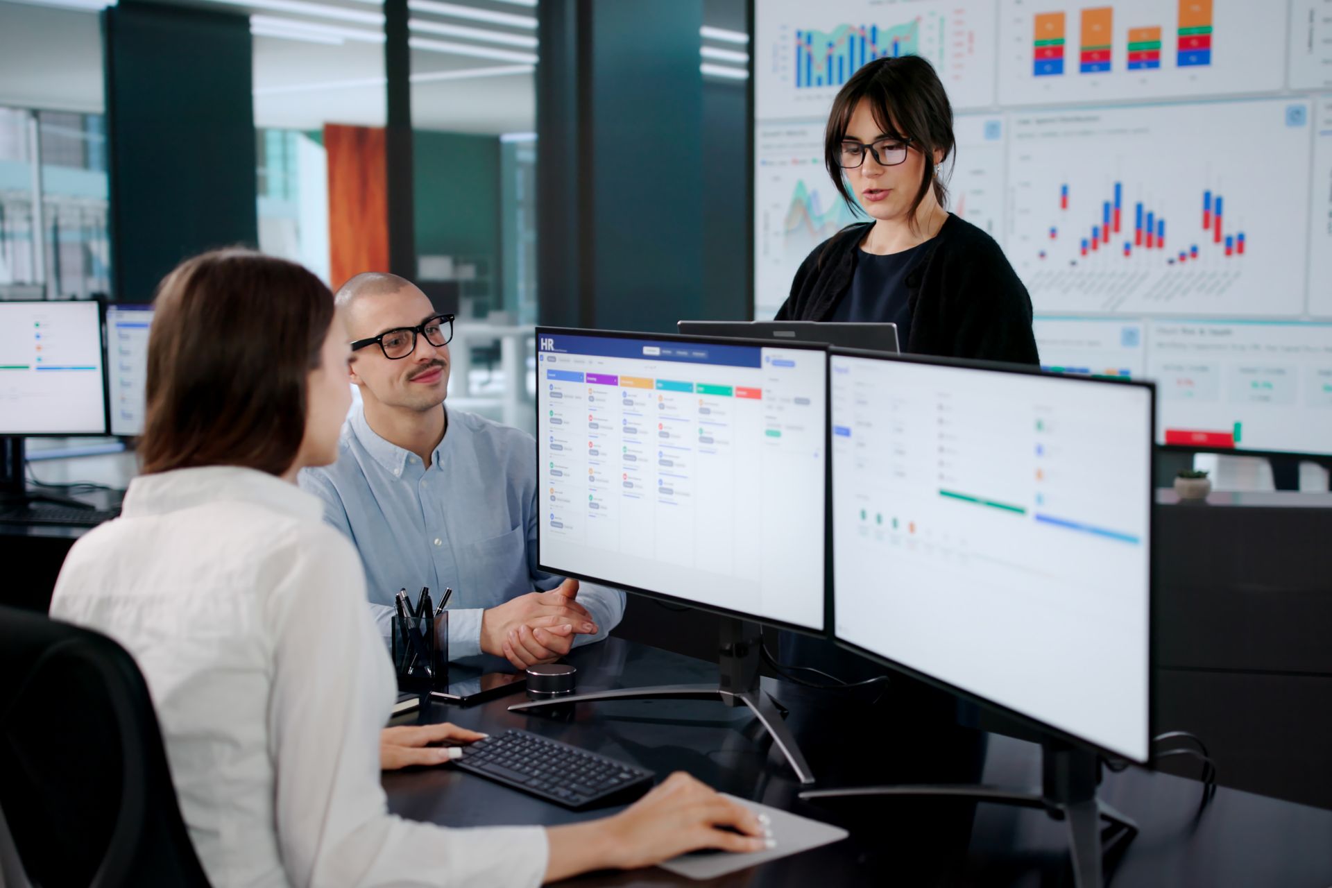 Three people collaborating around computer monitors displaying data visualizations in a modern office.