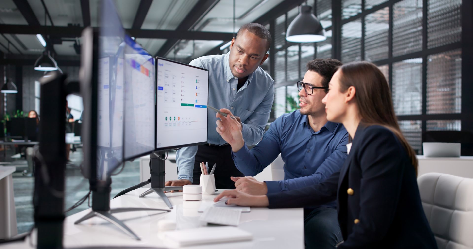 Three people in an office looking at computer screens; a man points at data, others watch.