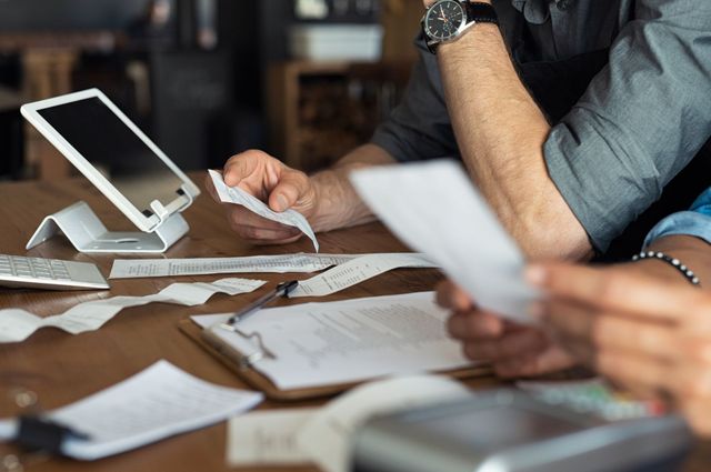 Woman in white shirt using a calculator, looking at documents with receipts and a laptop.