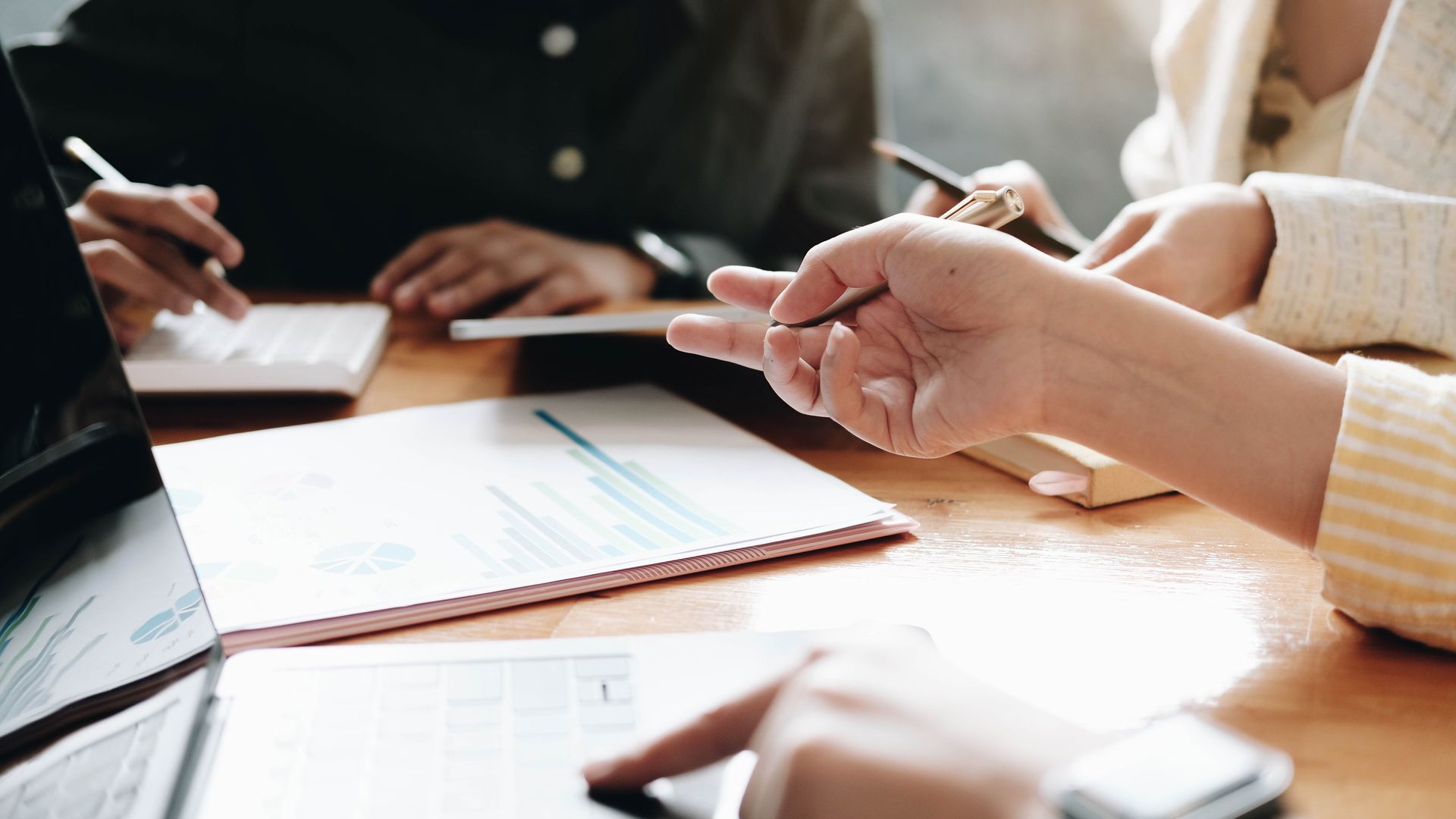 Hands pointing at papers and a laptop during a meeting. People are discussing finances.