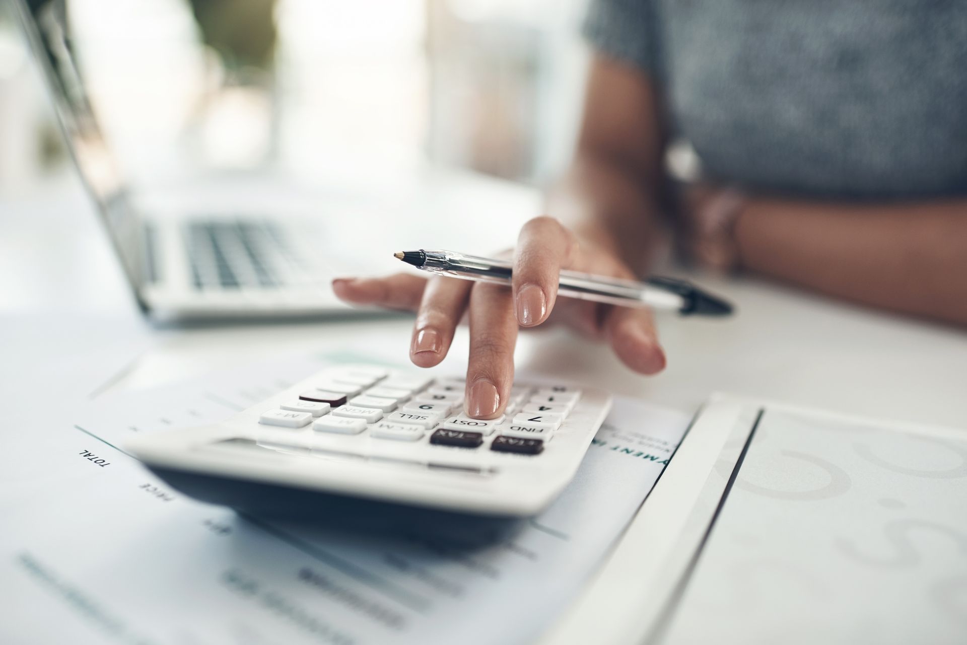 Person using a calculator with a pen, laptop and papers on a desk.
