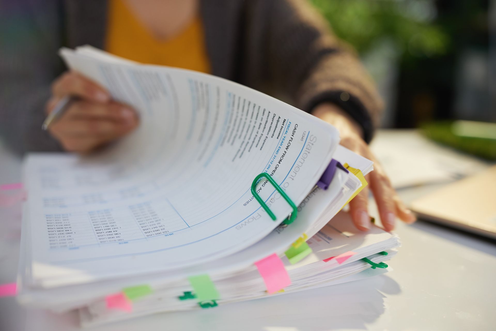 Person holding and flipping through a stack of papers with colorful sticky notes and a green paperclip.