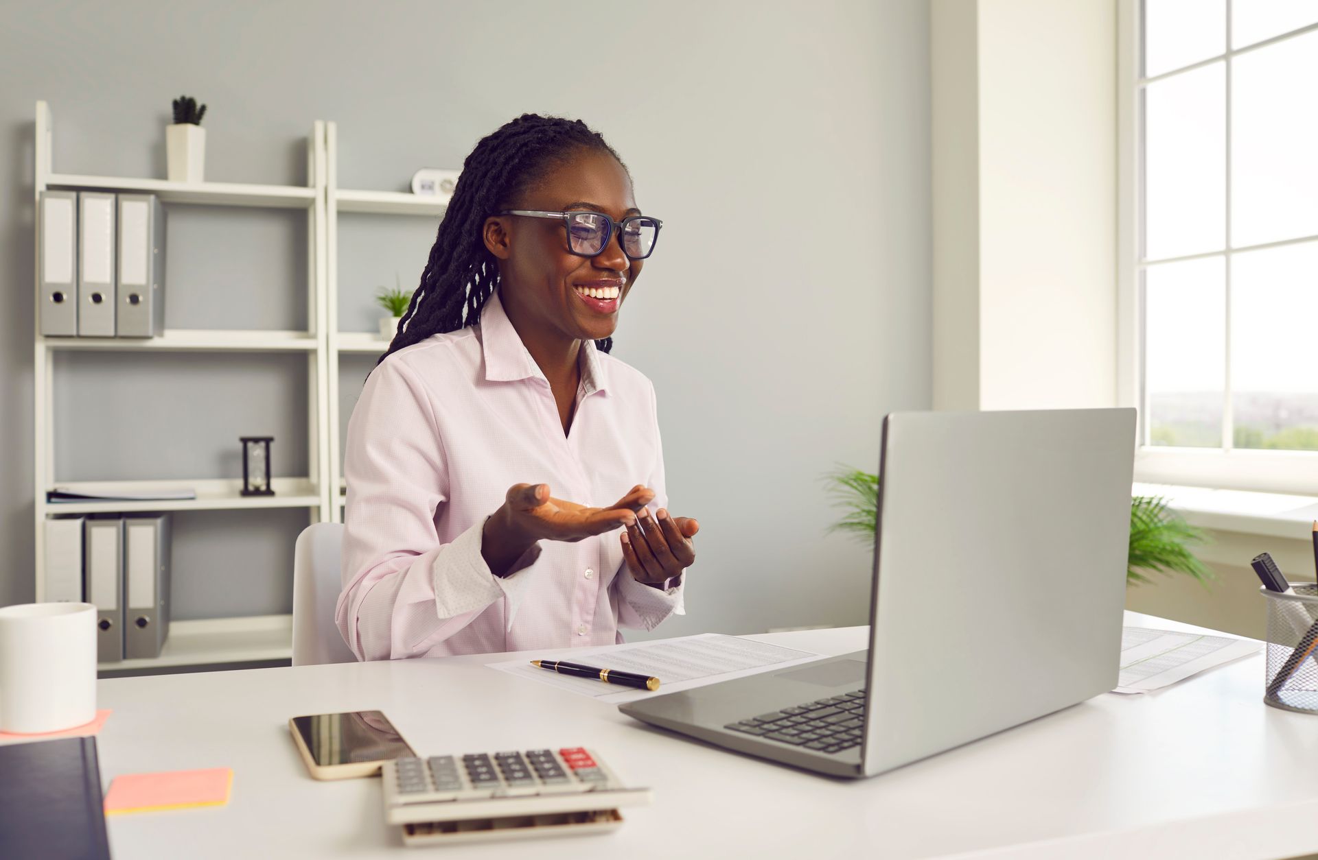 Woman with glasses smiles while video conferencing on a laptop at a desk in an office setting.