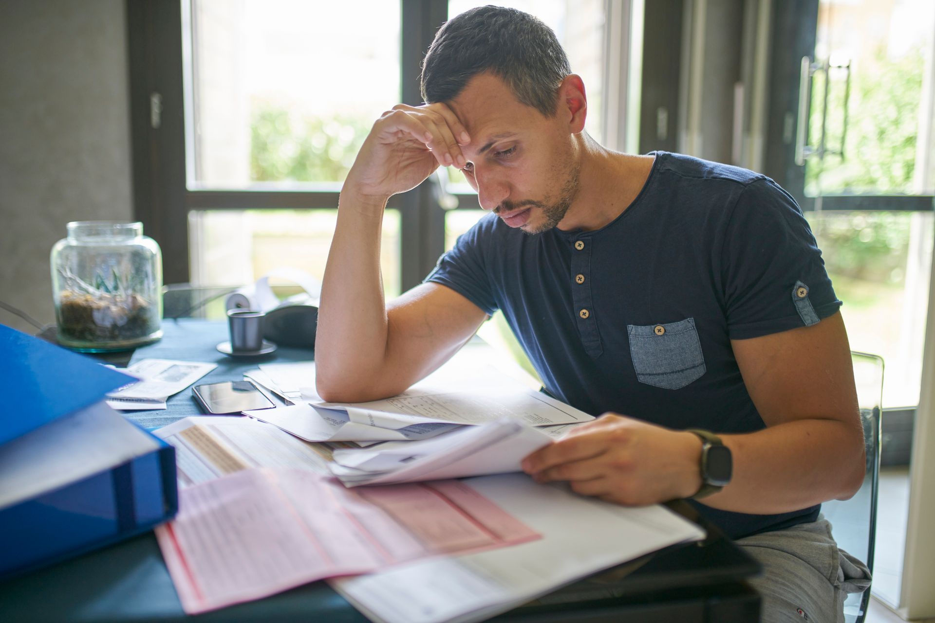 Man looking stressed, hand on forehead, reviewing paperwork at a table.