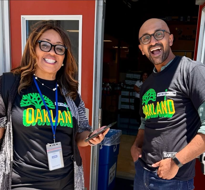Two smiling people in Oakland shirts standing in a doorway, one holding a phone