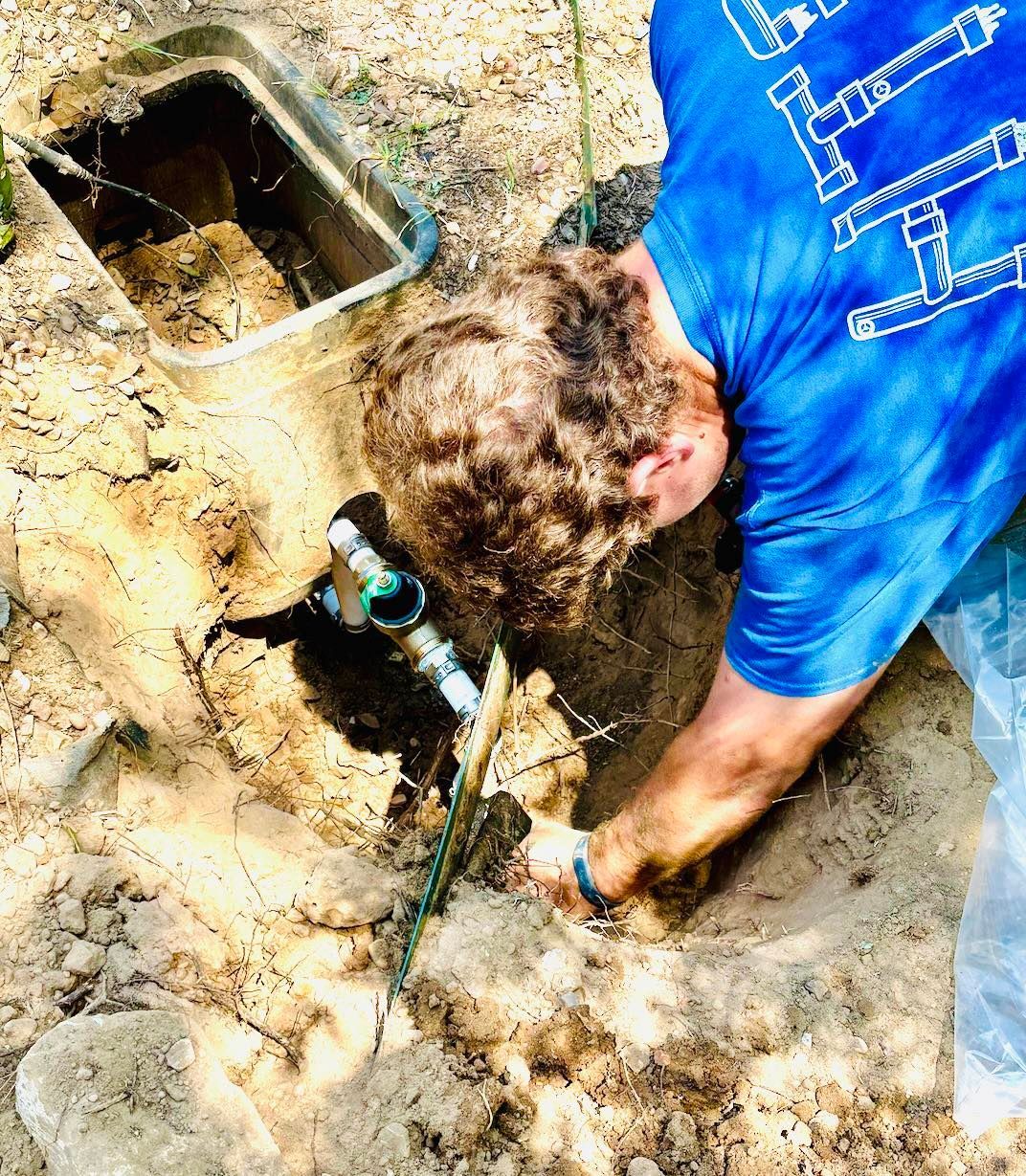 Man in blue shirt, working on underground pipe in dirt near an open valve box.