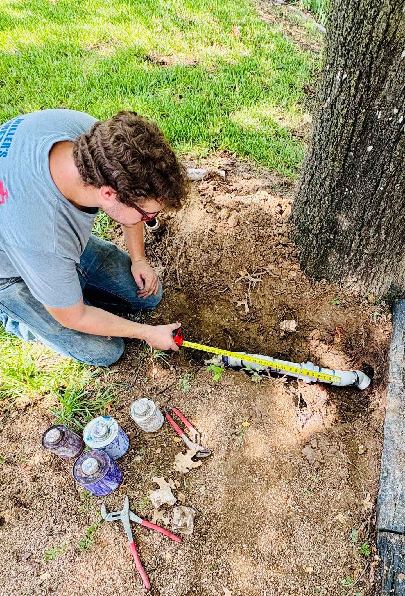 Person measuring ground near a tree with a tape measure, tools and flowers nearby.