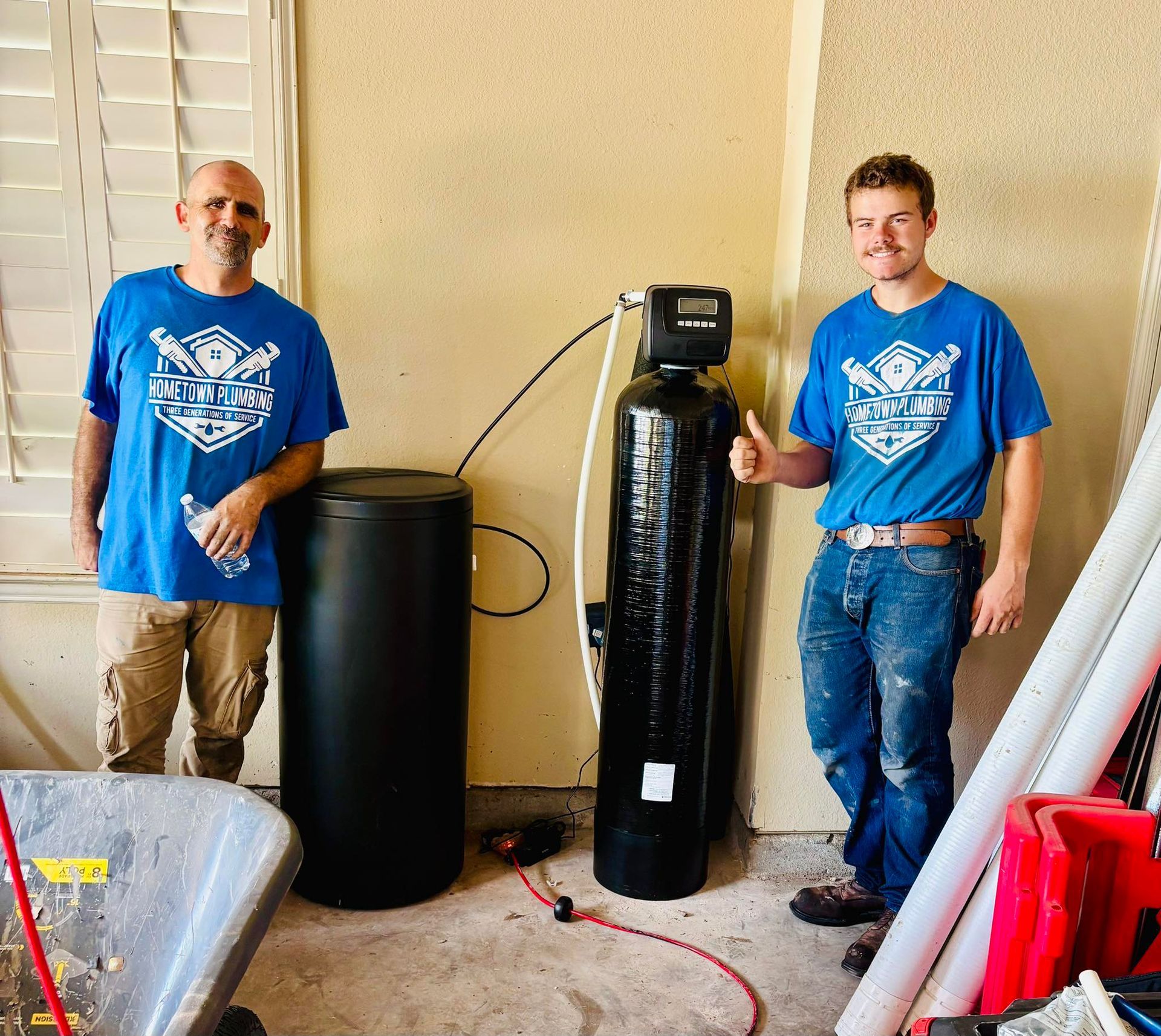 Two men stand next to a water filtration system. They are wearing blue shirts and smiling.