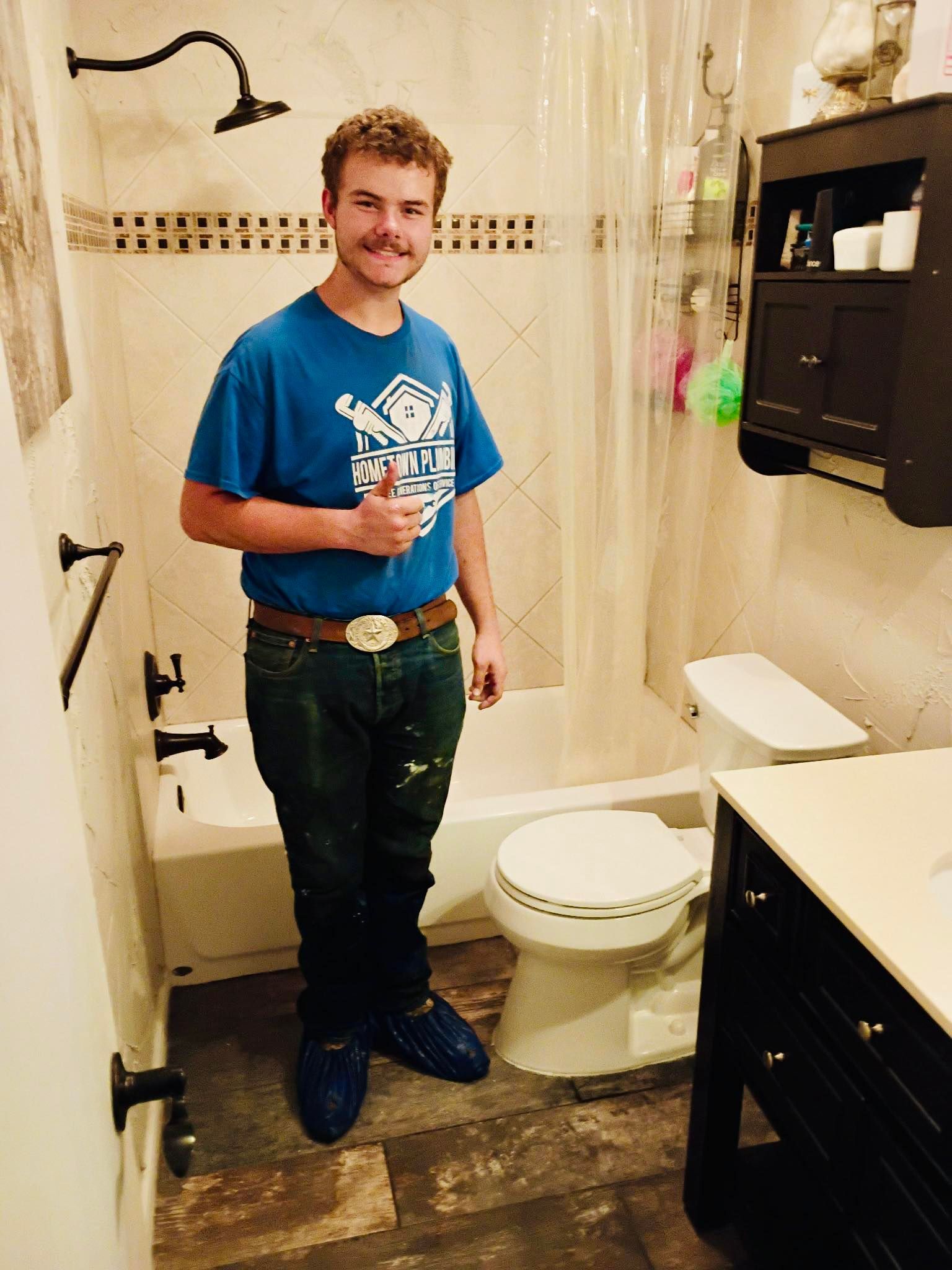 Man in blue shirt, jeans, and work boots gives thumbs up in a newly renovated bathroom with new fixtures and tile.