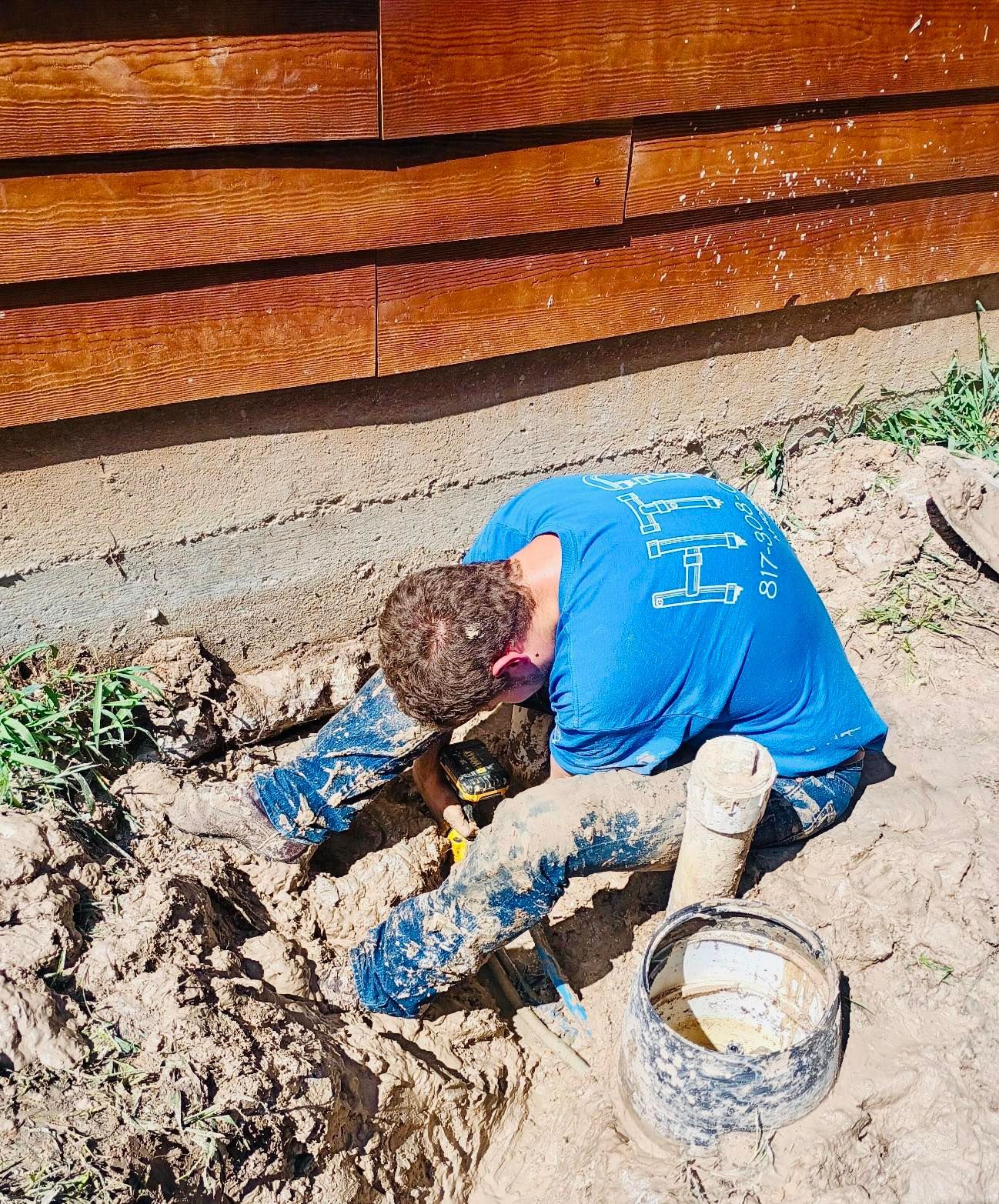Person in blue shirt working on plumbing in a ditch near a building.