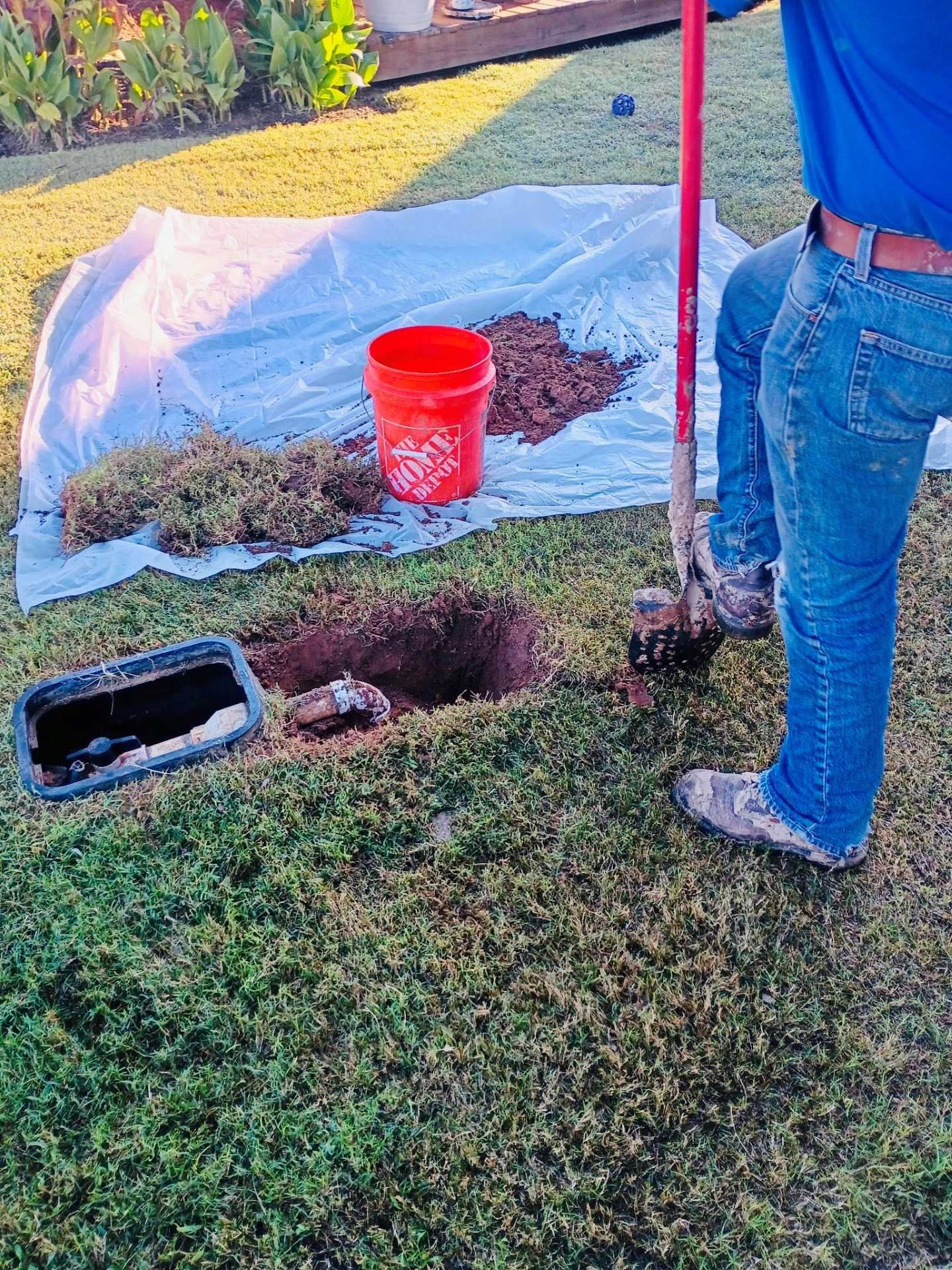 Person digging in a yard near a water meter box; dirt pile, red bucket, and tarp are present.
