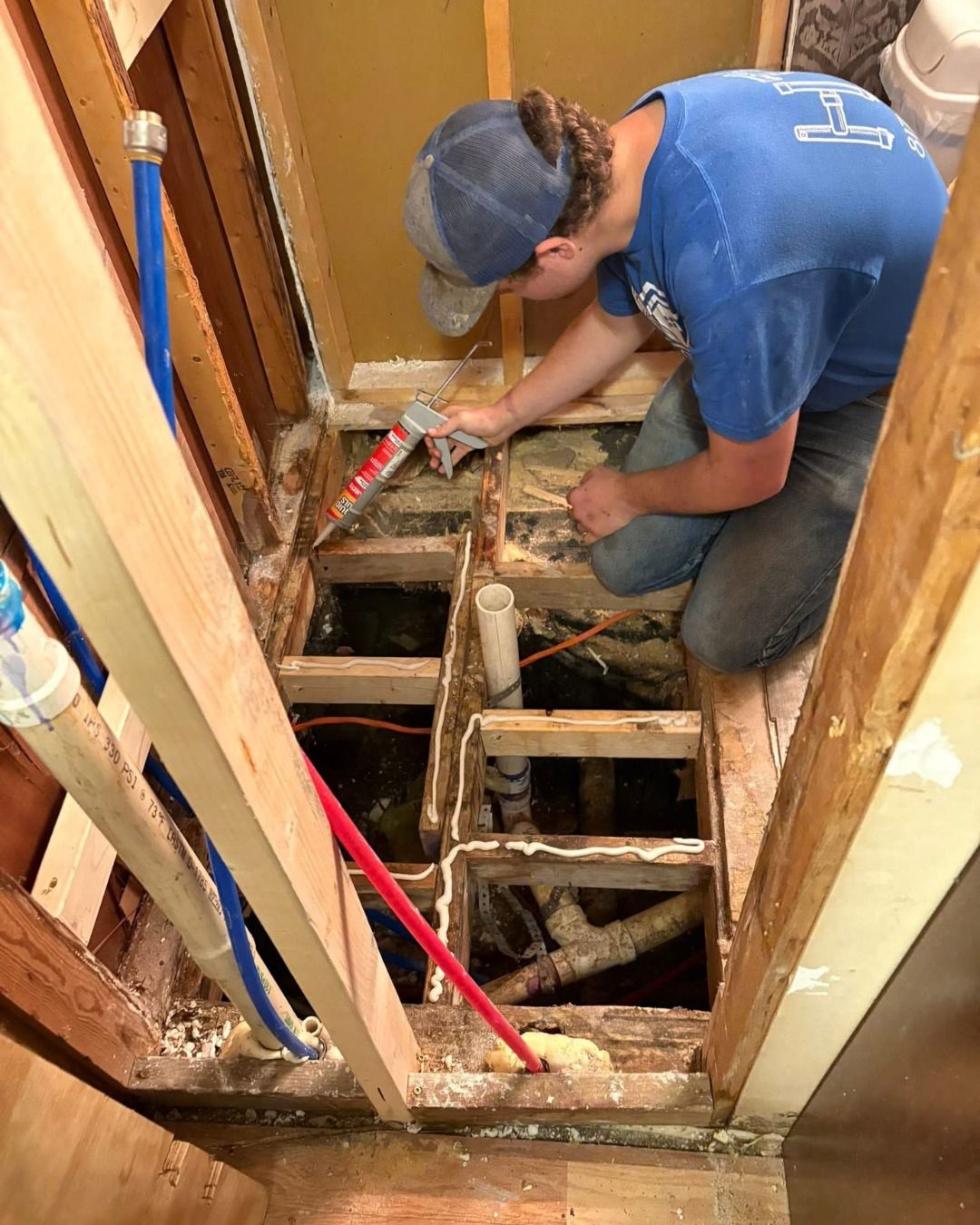 Person applying sealant to a bathroom floor during construction; exposed plumbing visible.