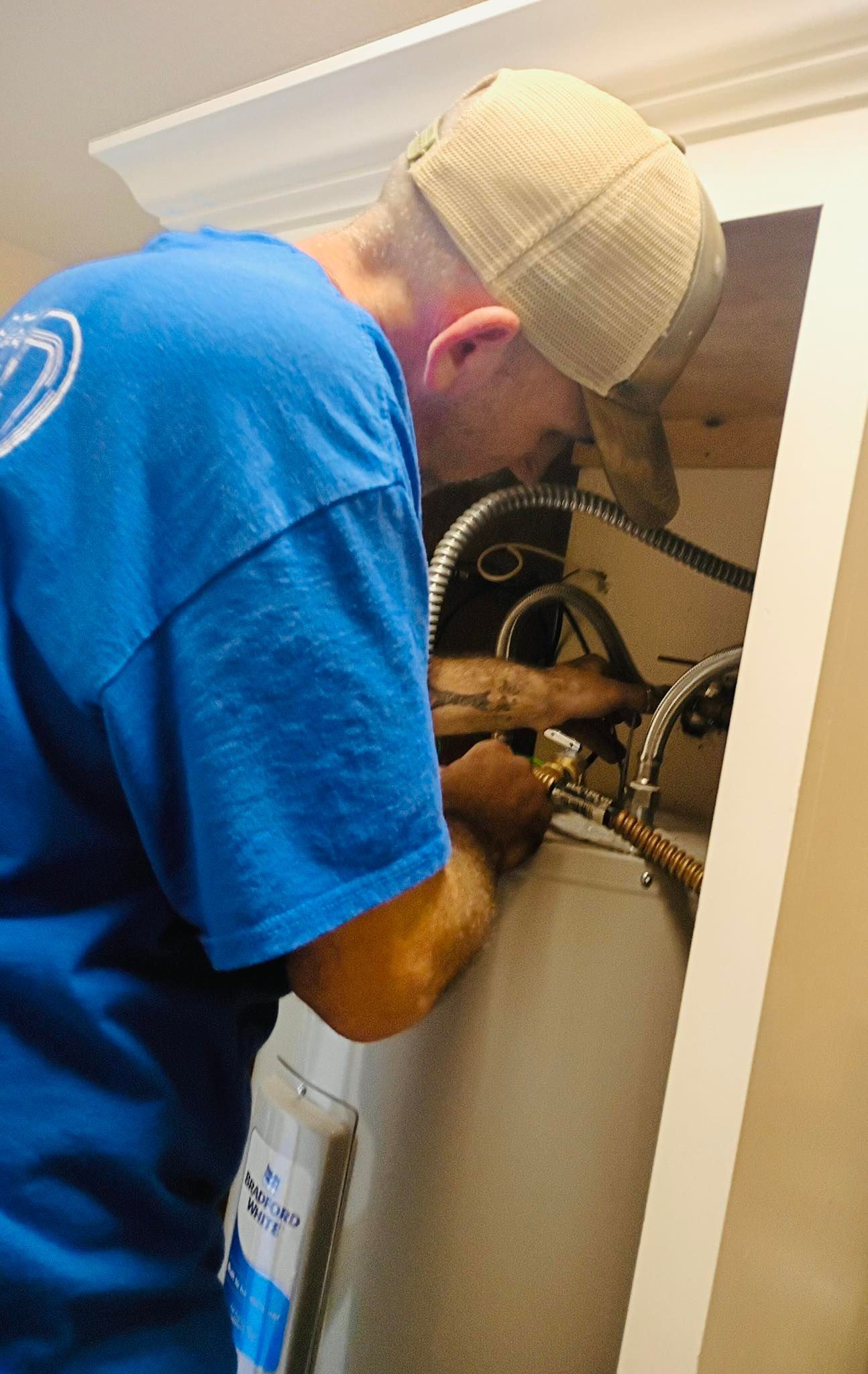 Plumber in blue shirt and baseball cap working on a water heater, located in a cabinet.