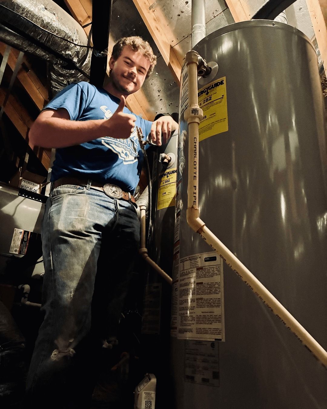 Man in blue shirt gives thumbs up next to a water heater in an attic, with various pipes visible.