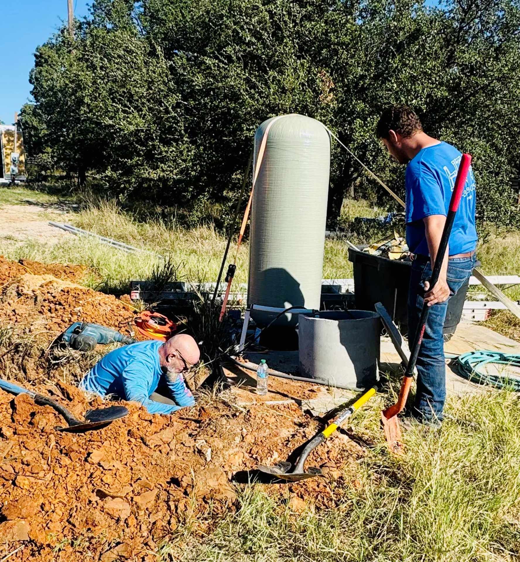 Two people working on outdoor equipment near a well. One in a hole, one holding a shovel.