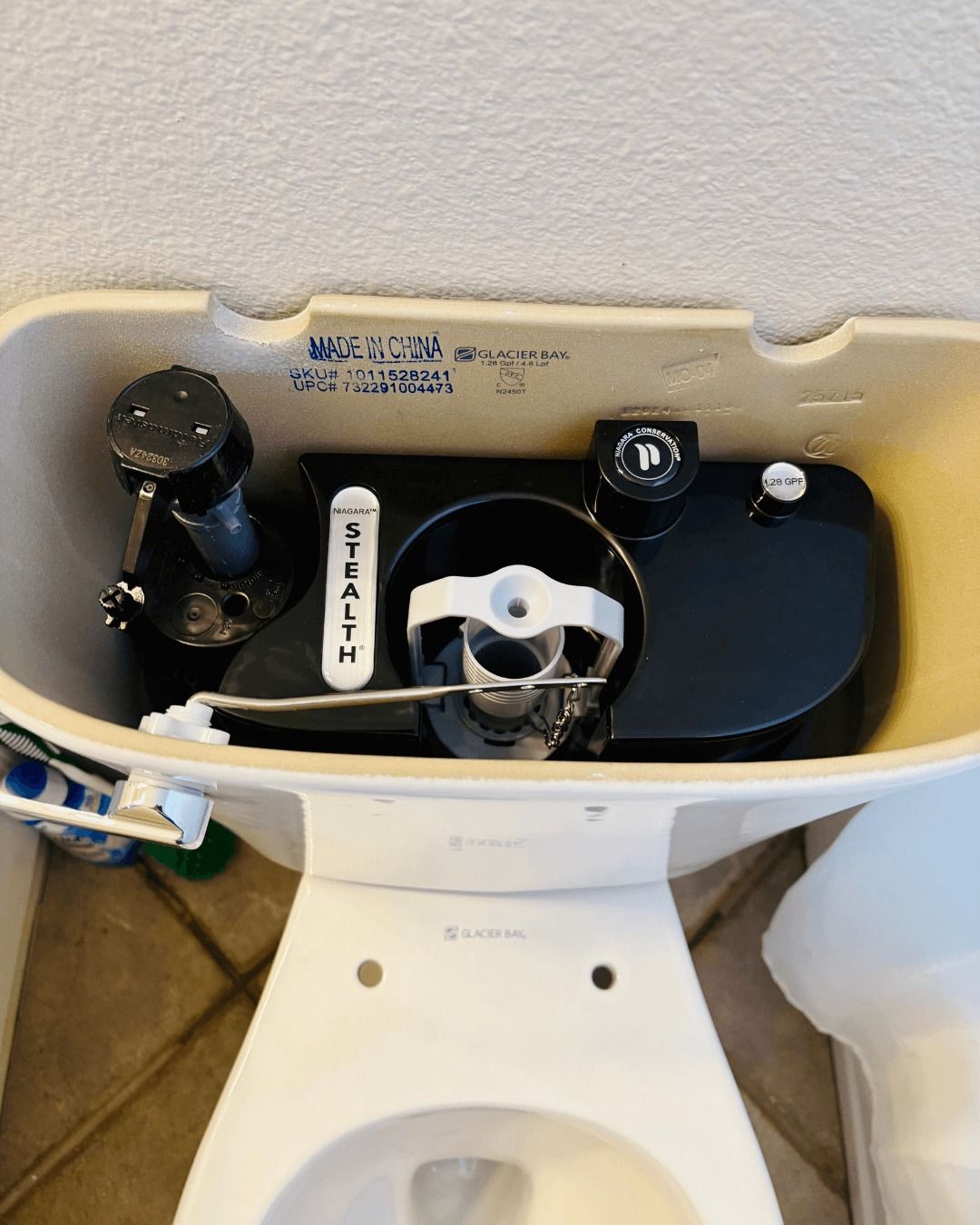 Toilet tank interior showing black fill valve, flush valve, and other components. Beige tank, white components.
