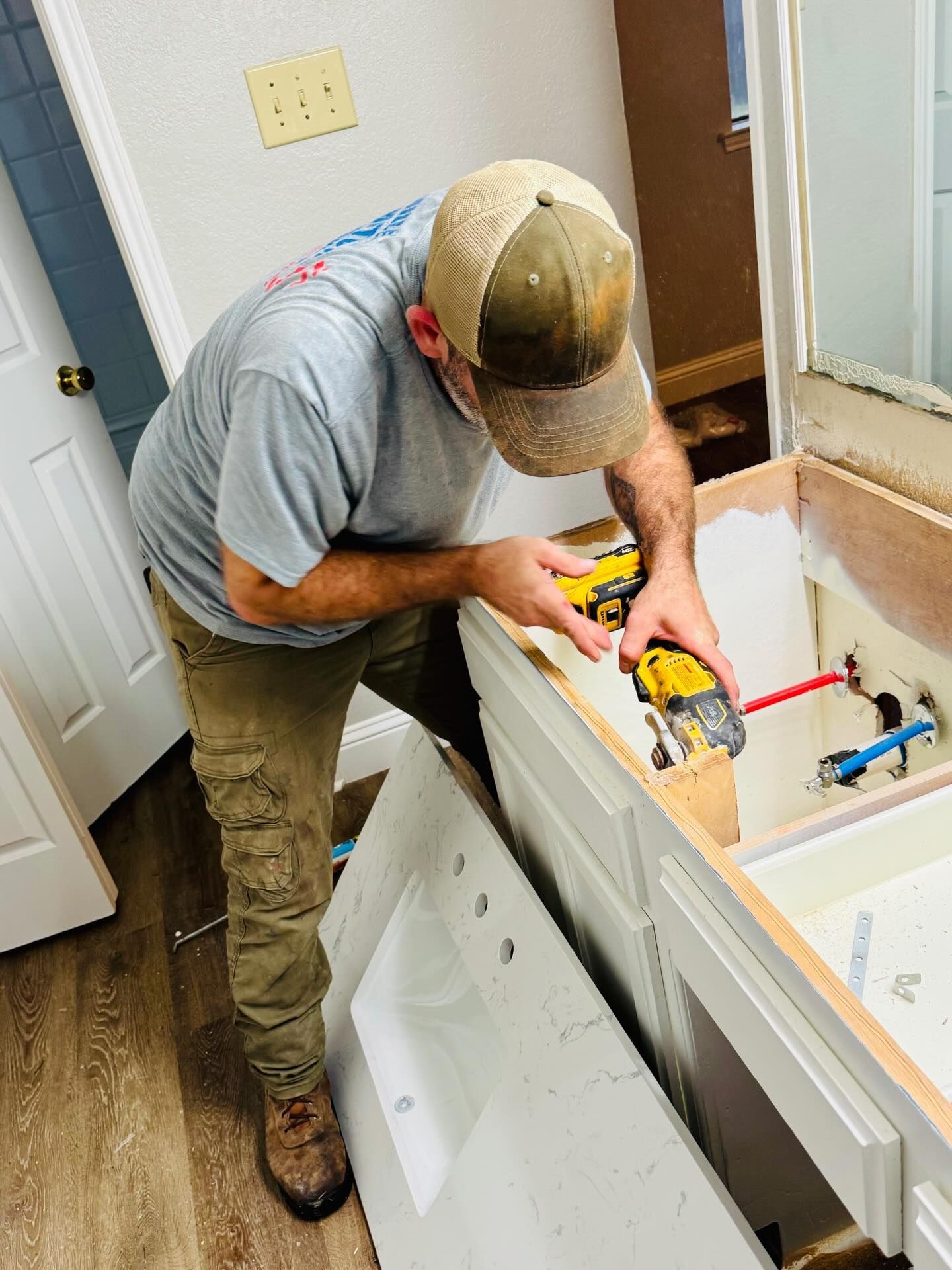 Person in a cap and work clothes using a power tool to work on a bathroom vanity with a sink.