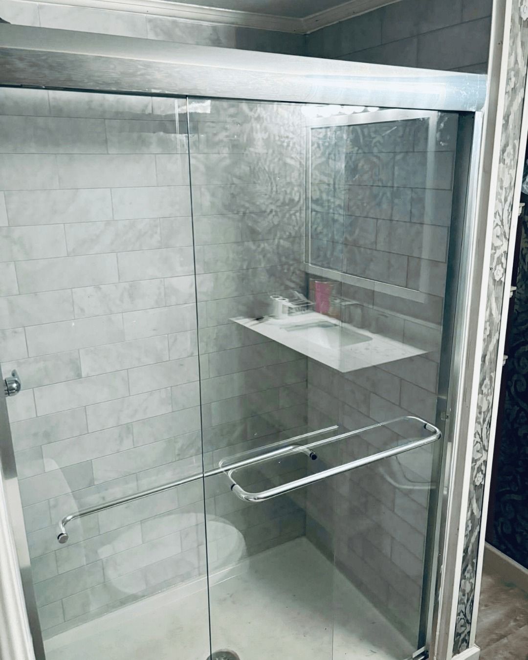 Shower with glass doors and a white marble-like tile background, featuring a towel rack and shelf.