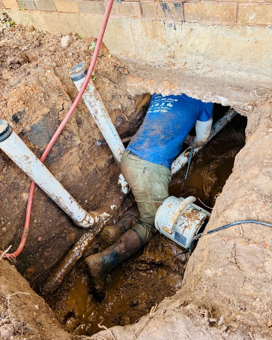 Plumber in a trench, repairing pipes. Muddy, wearing blue shirt, and reaching into the hole.