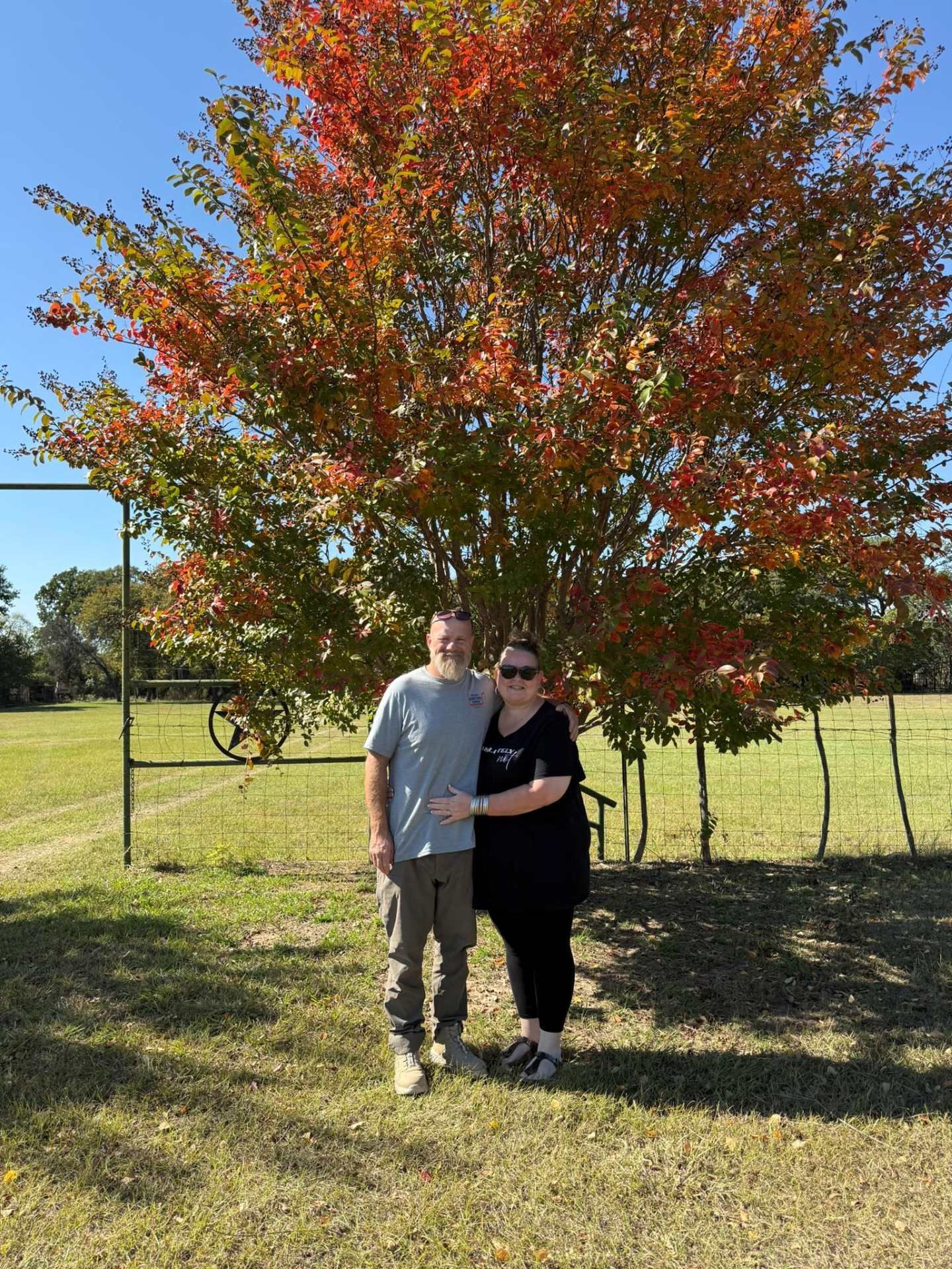 Couple standing under a tree with red and orange leaves; outdoors on a sunny day.