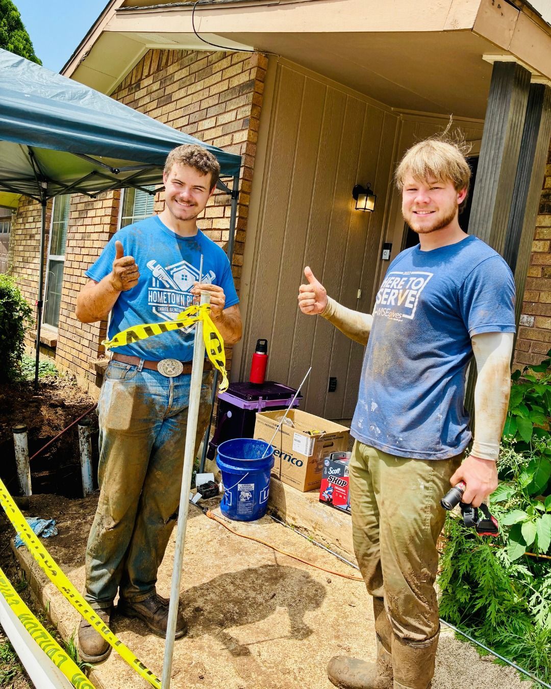 Two men covered in mud give thumbs up next to a house under construction.