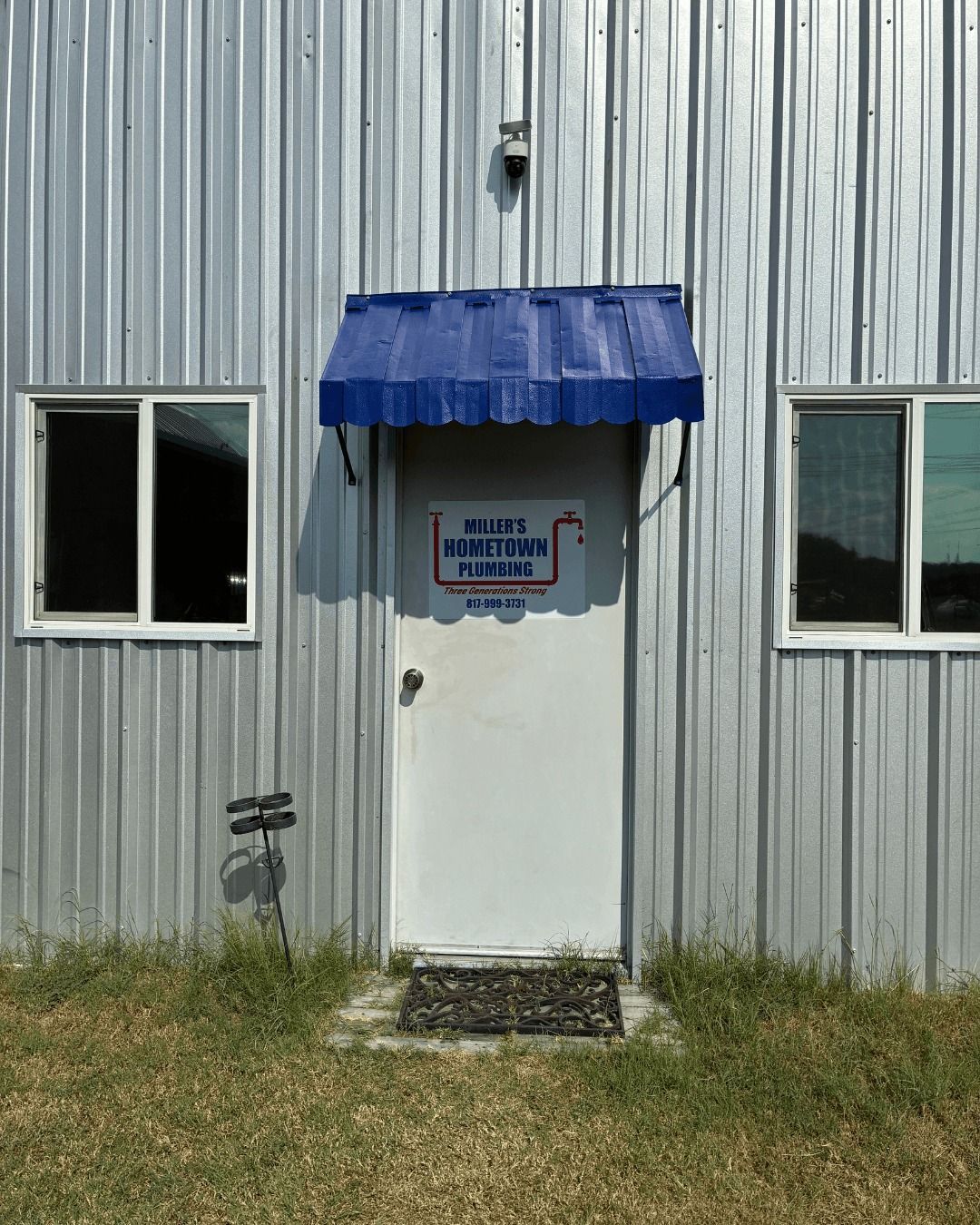 White door with blue awning on a corrugated metal building, flanked by windows. 
