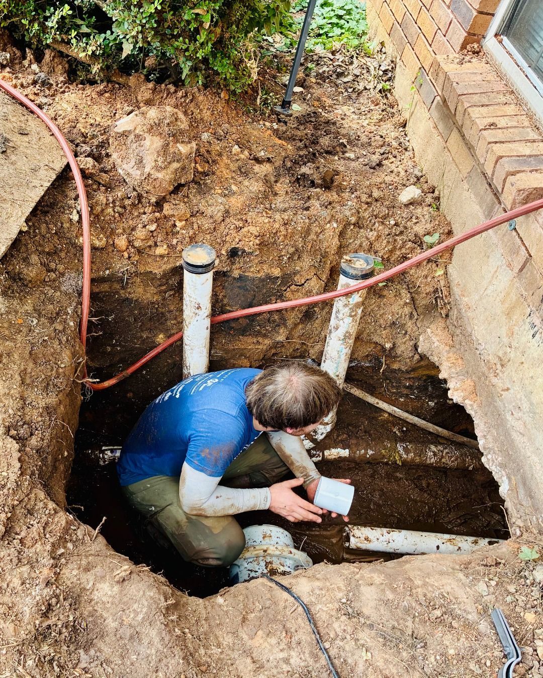 A person in a hole inspecting pipes next to a brick building.
