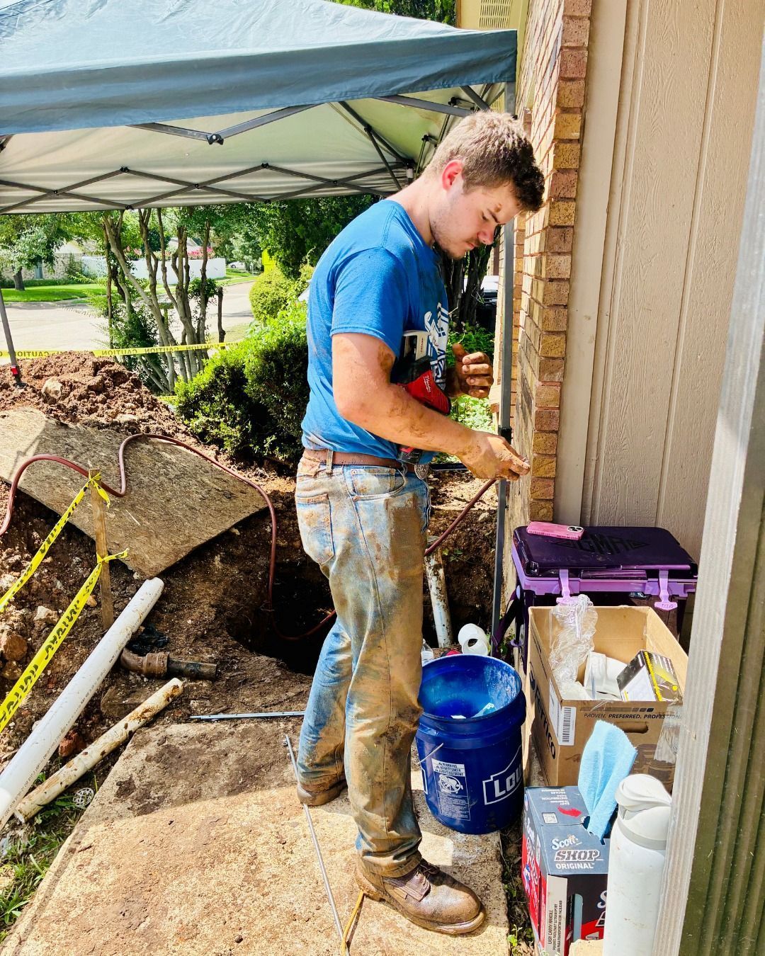 Man in blue shirt and muddy jeans works on pipes beside a building, near a trench.