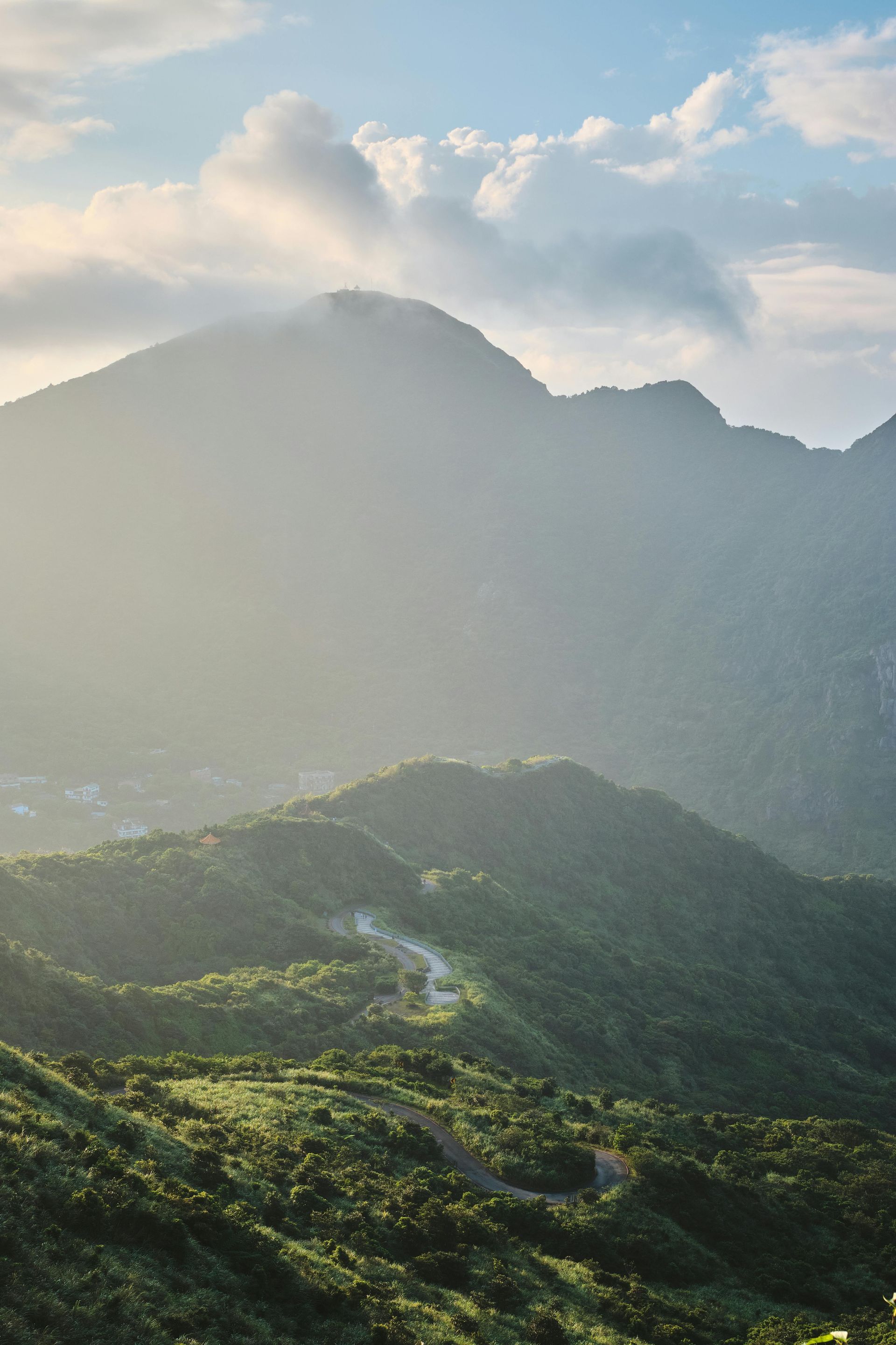 Green mountains and winding road under a cloudy, hazy sky.